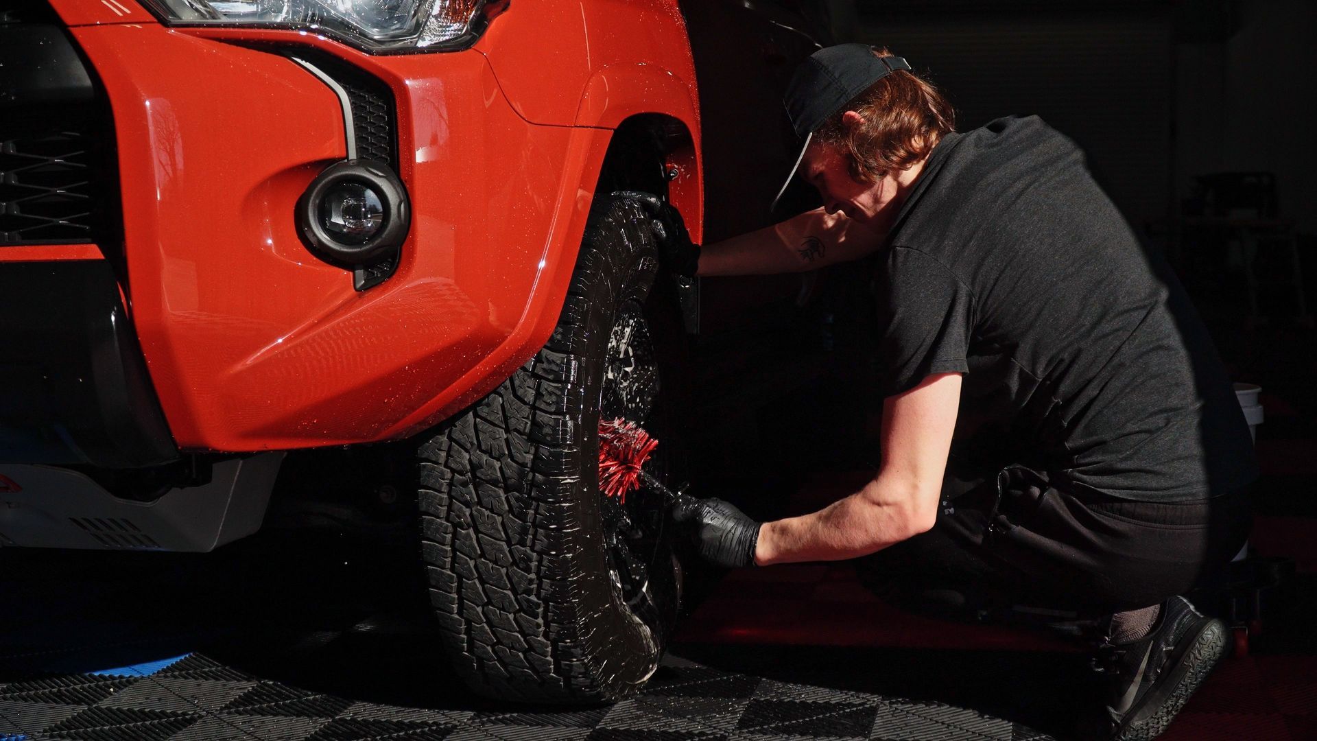 Mechanic in black clothing works under a red car's front wheel in a dark garage