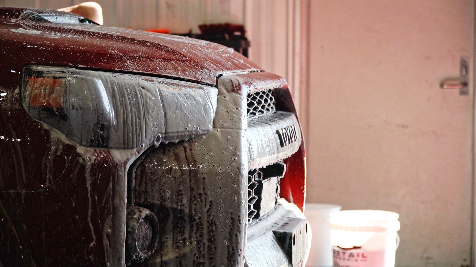 Red tractor front covered in suds during a car wash, with a bucket in the background.