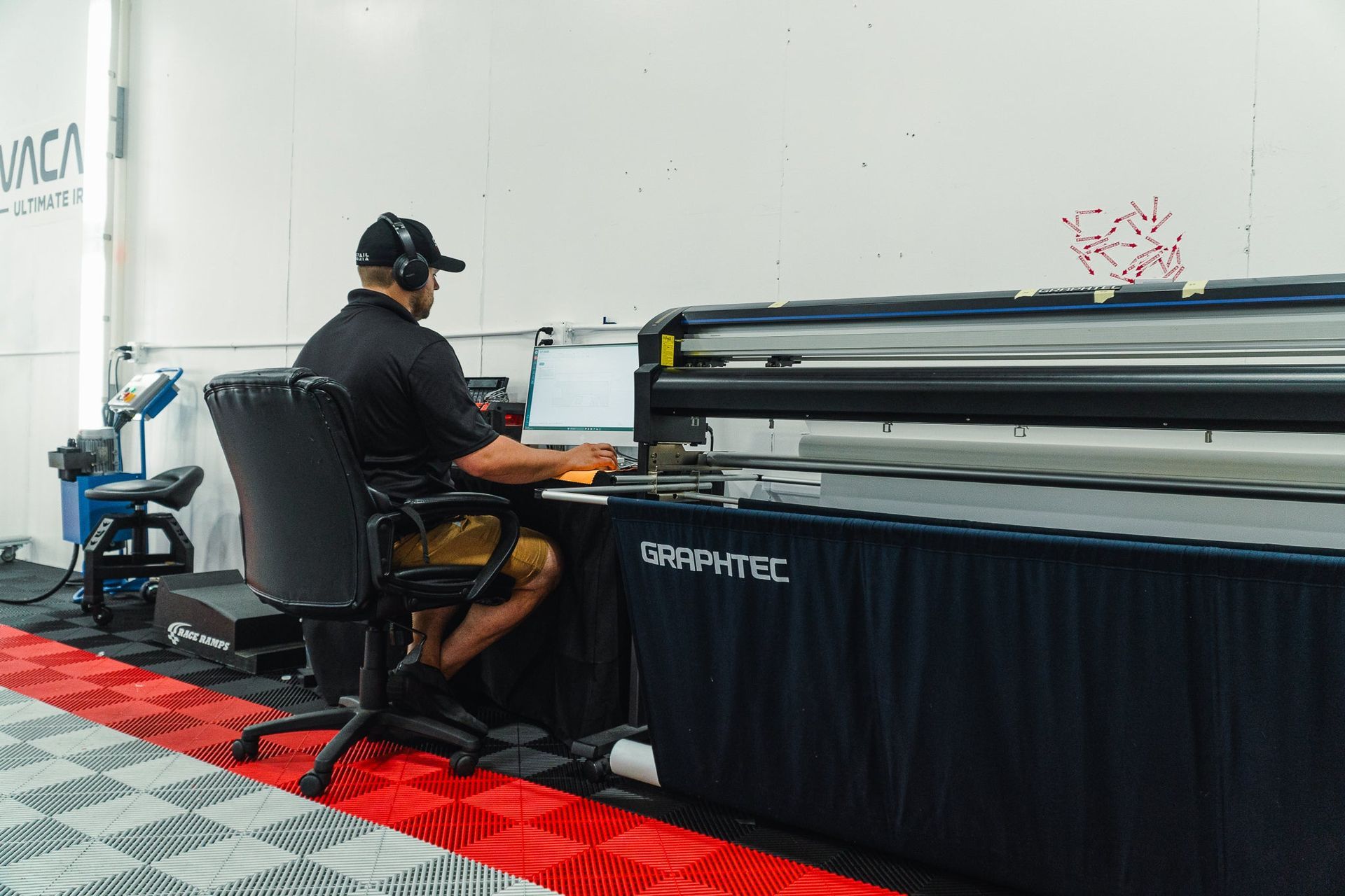 Person working at a large-format printer in a workshop, with a laptop on a desk.