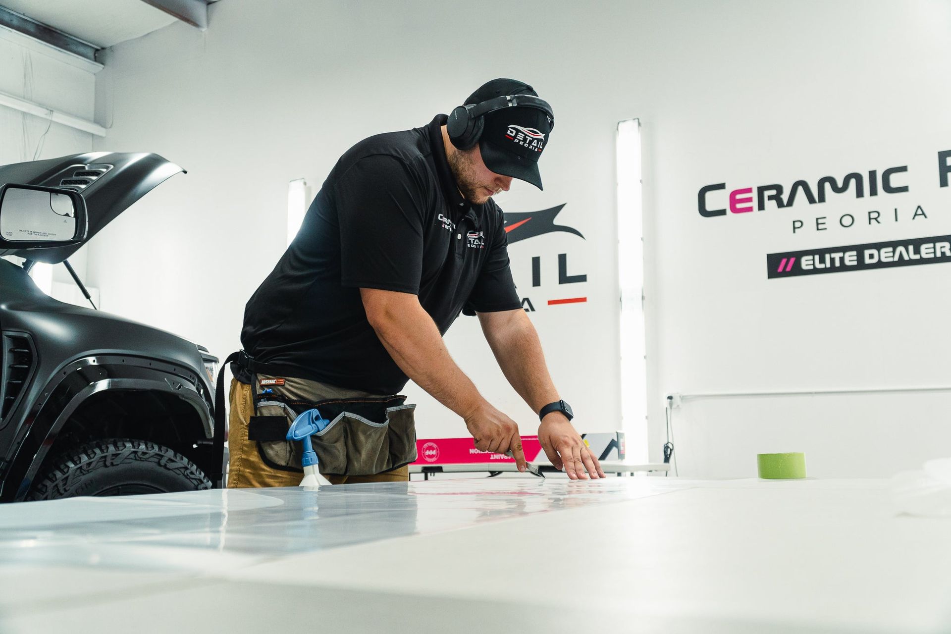 Detailer polishing a white car in a ceramic coating shop, with a black vehicle nearby and signage in the background