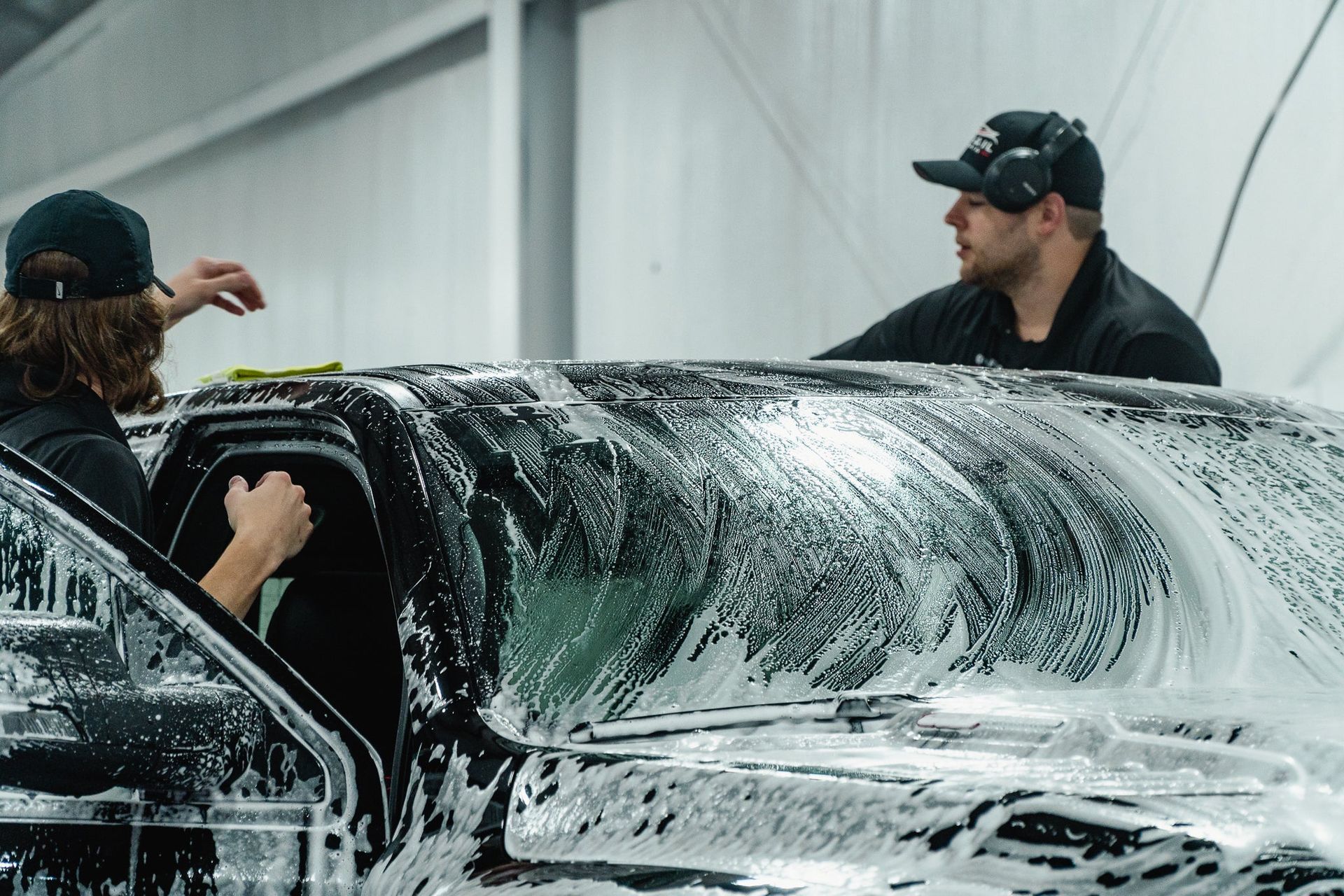 Two men washing a black car covered in white foam in a garage