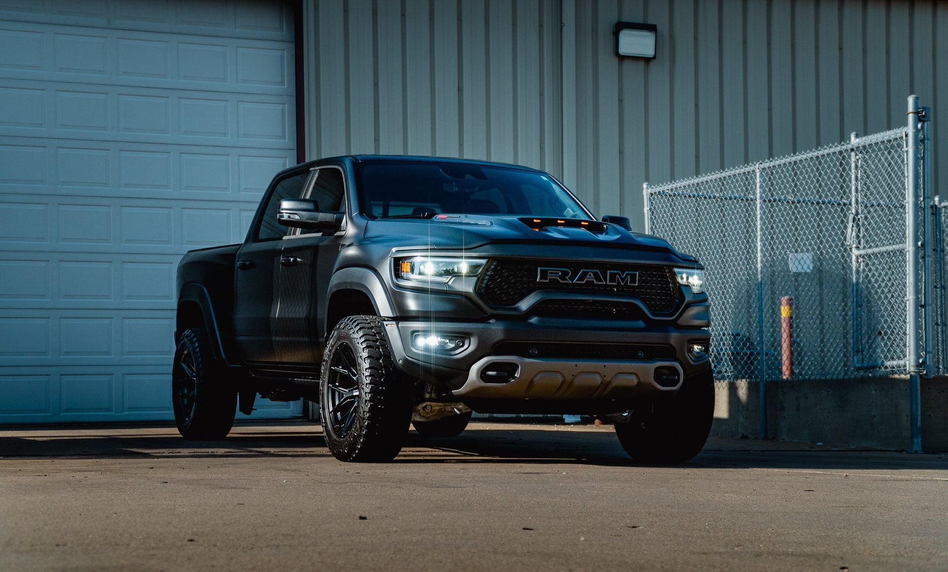 Dark gray pickup truck parked outside a warehouse beside a chain-link fence.