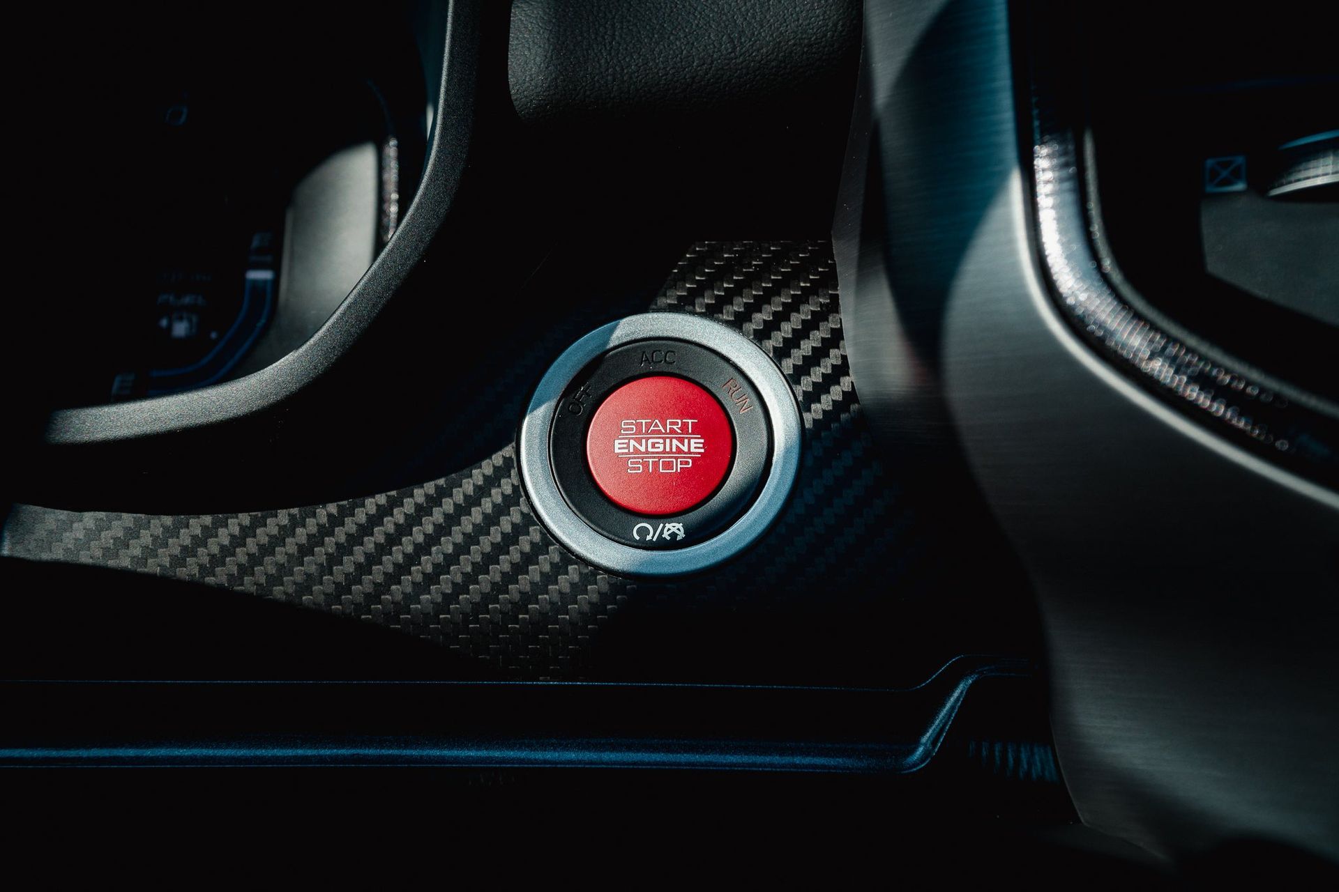 Close-up of a red sport car ignition button on a carbon-fiber dashboard.