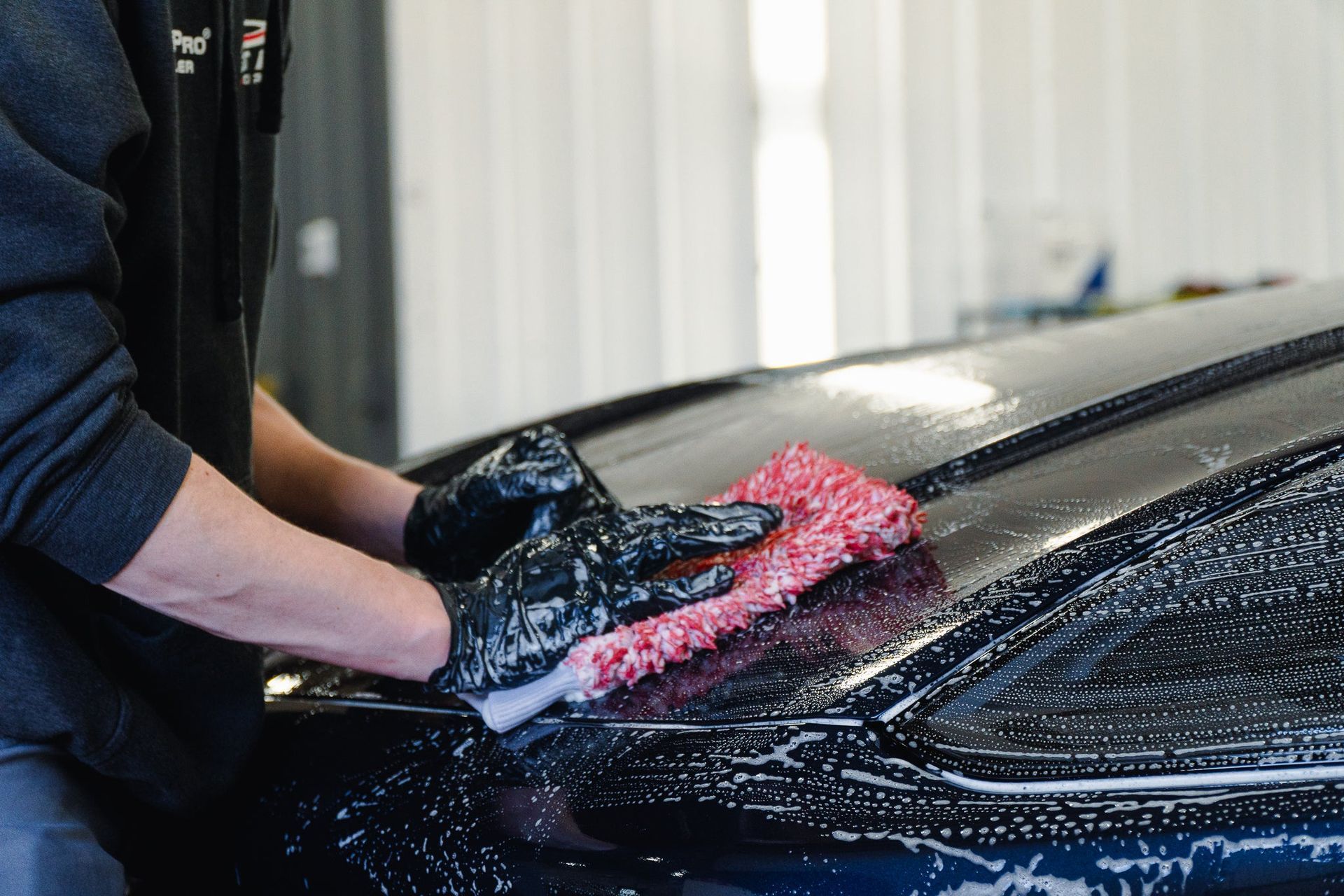 Person washing a black car hood with red foam and a sponge in a garage
