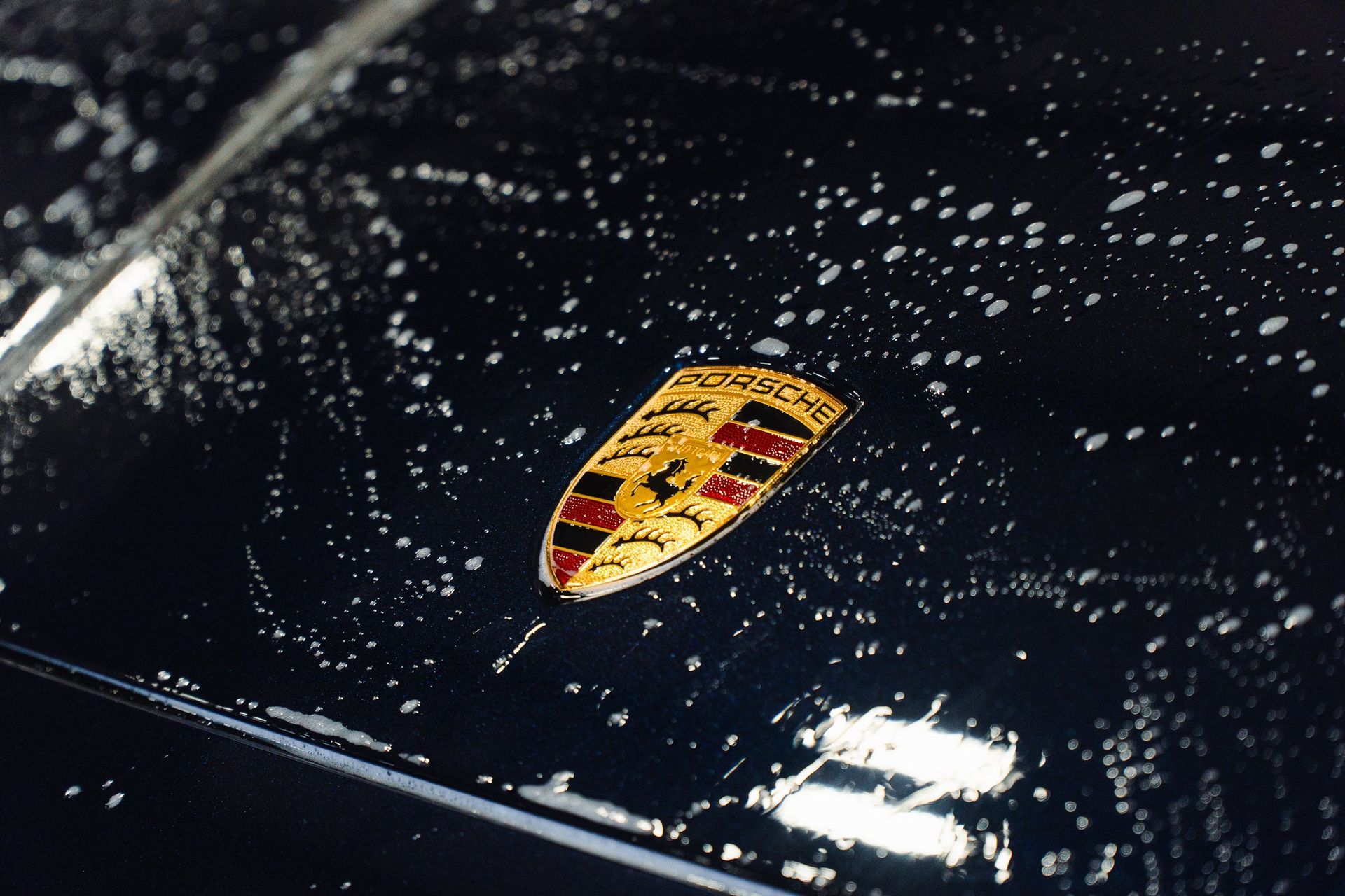 Porsche hood emblem on a glossy black car, speckled with water droplets