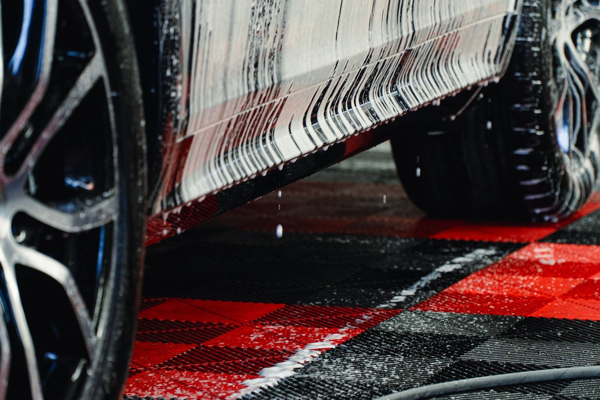 Close-up of a car being washed, with soapy water on black tires and red wet pavement