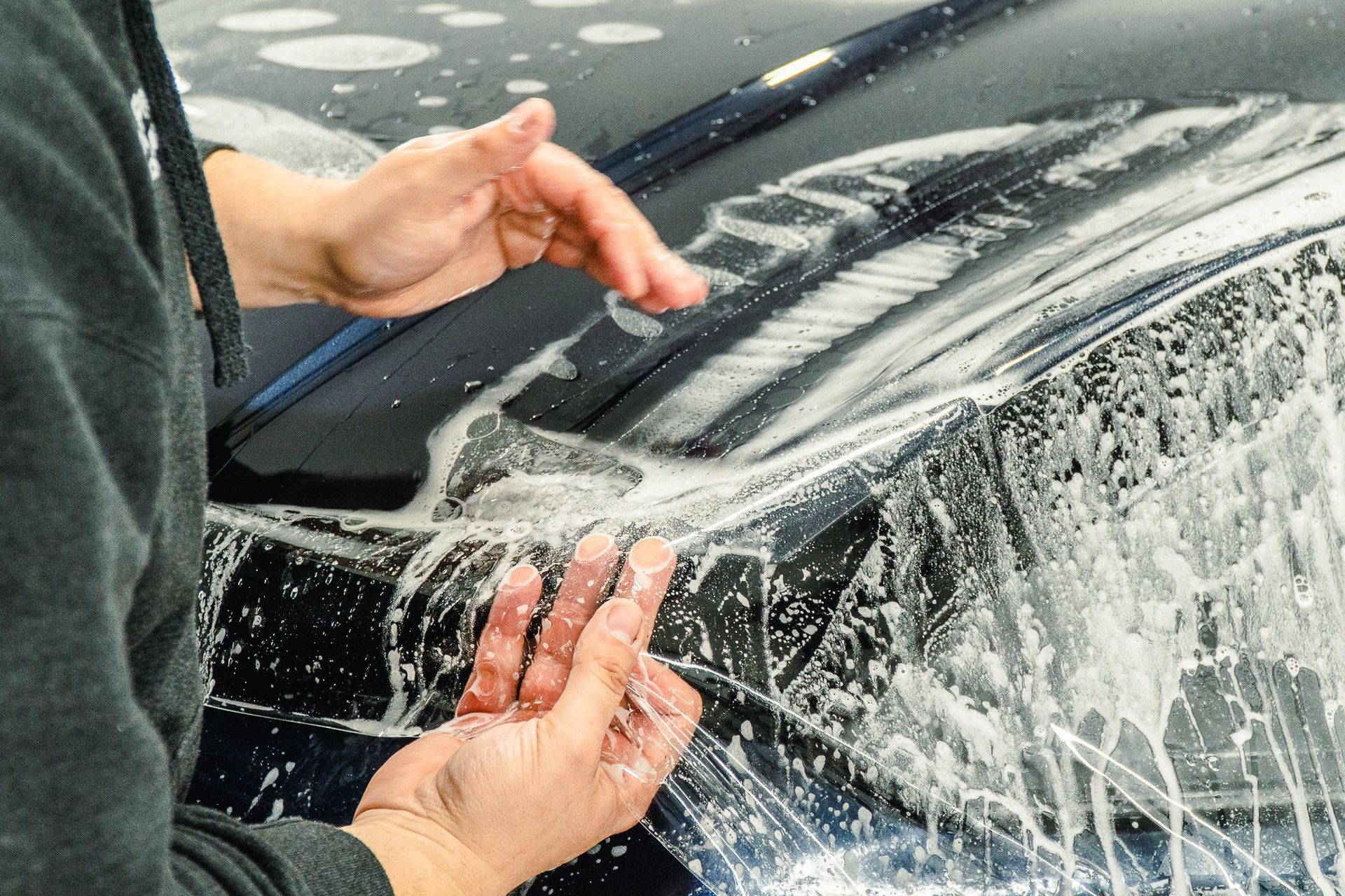 Hands washing a black car windshield with soap suds and a sponge