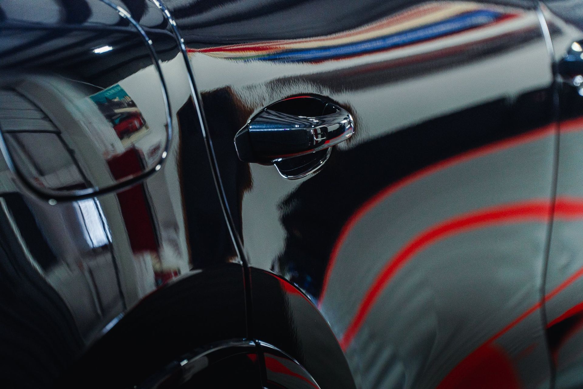 Close-up of a glossy black car door with chrome handle and red reflections on the body.