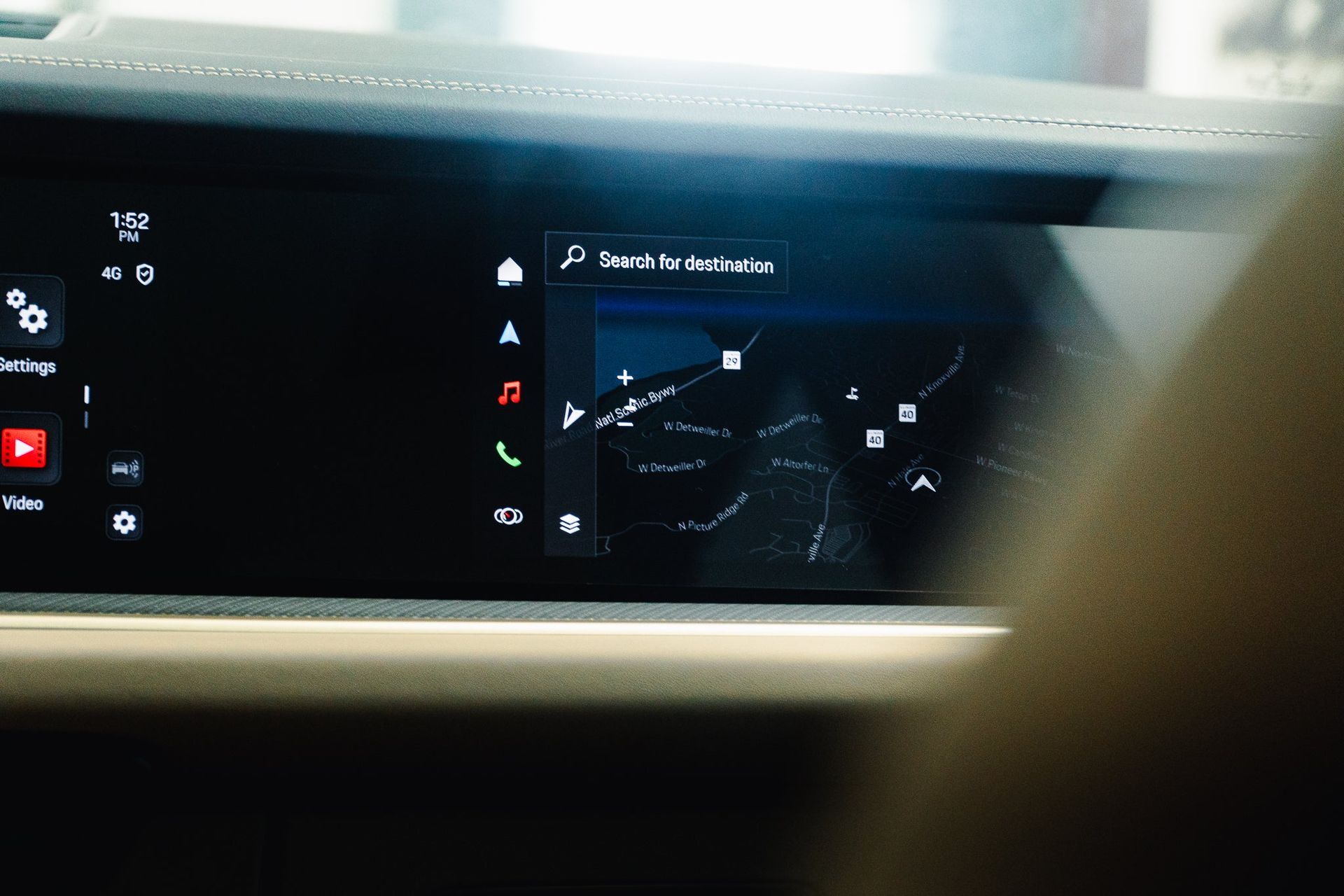 Car dashboard touchscreen showing navigation map and app icons, partially obscured by a hand