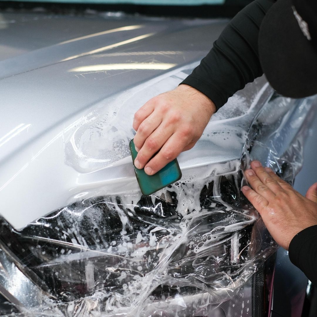 Person scrapes protective film off a silver car hood with a green tool