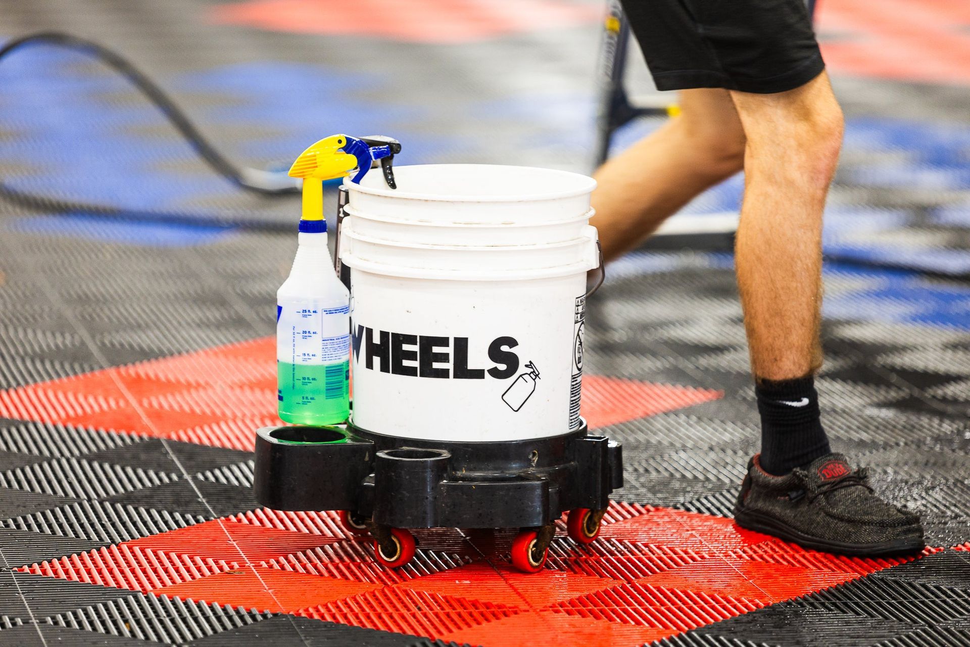 A wheel-mounted white bucket with spray bottles on a red-and-black sports court beside a person’s legs