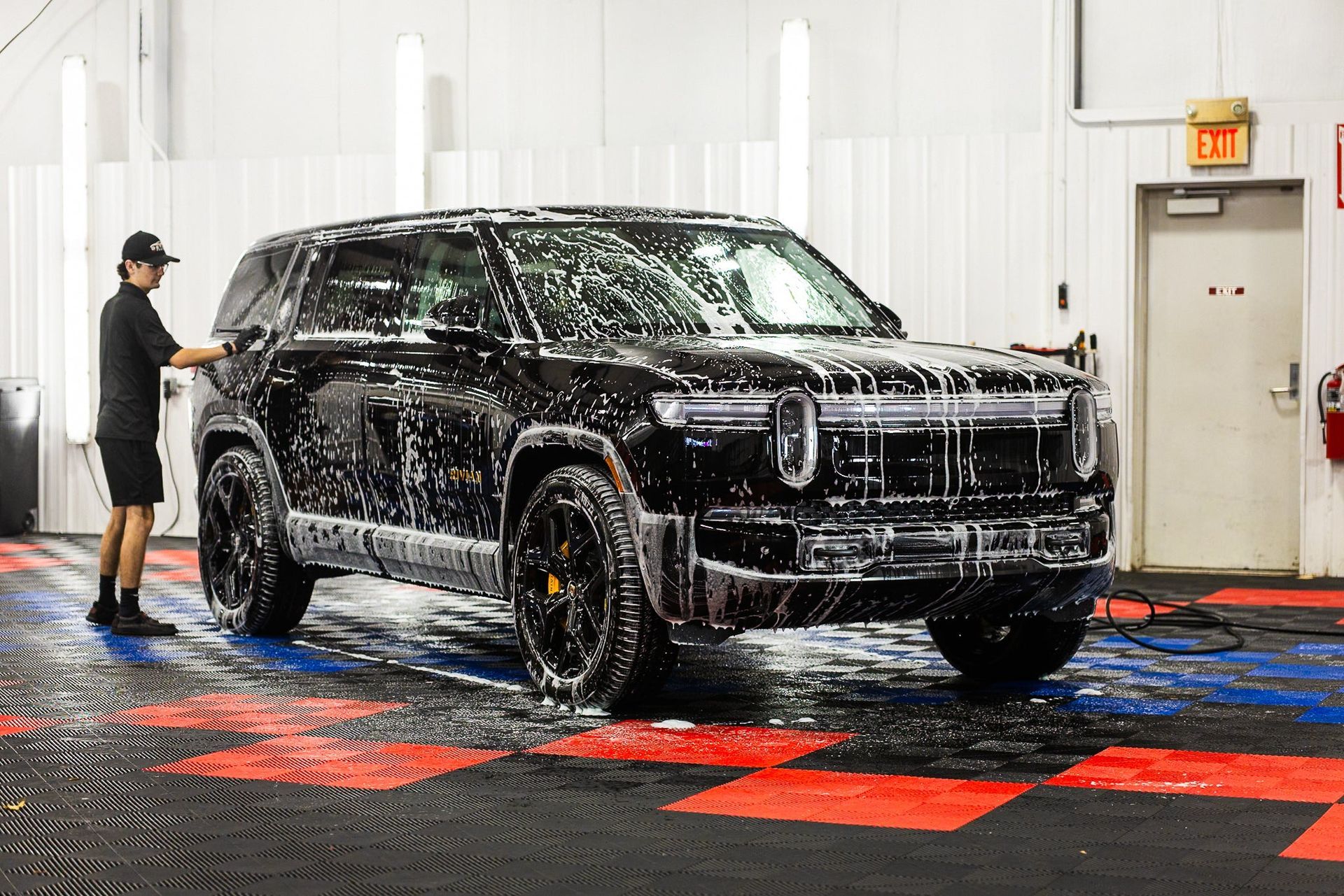 Black SUV being washed in a car wash bay, with an attendant spraying water.