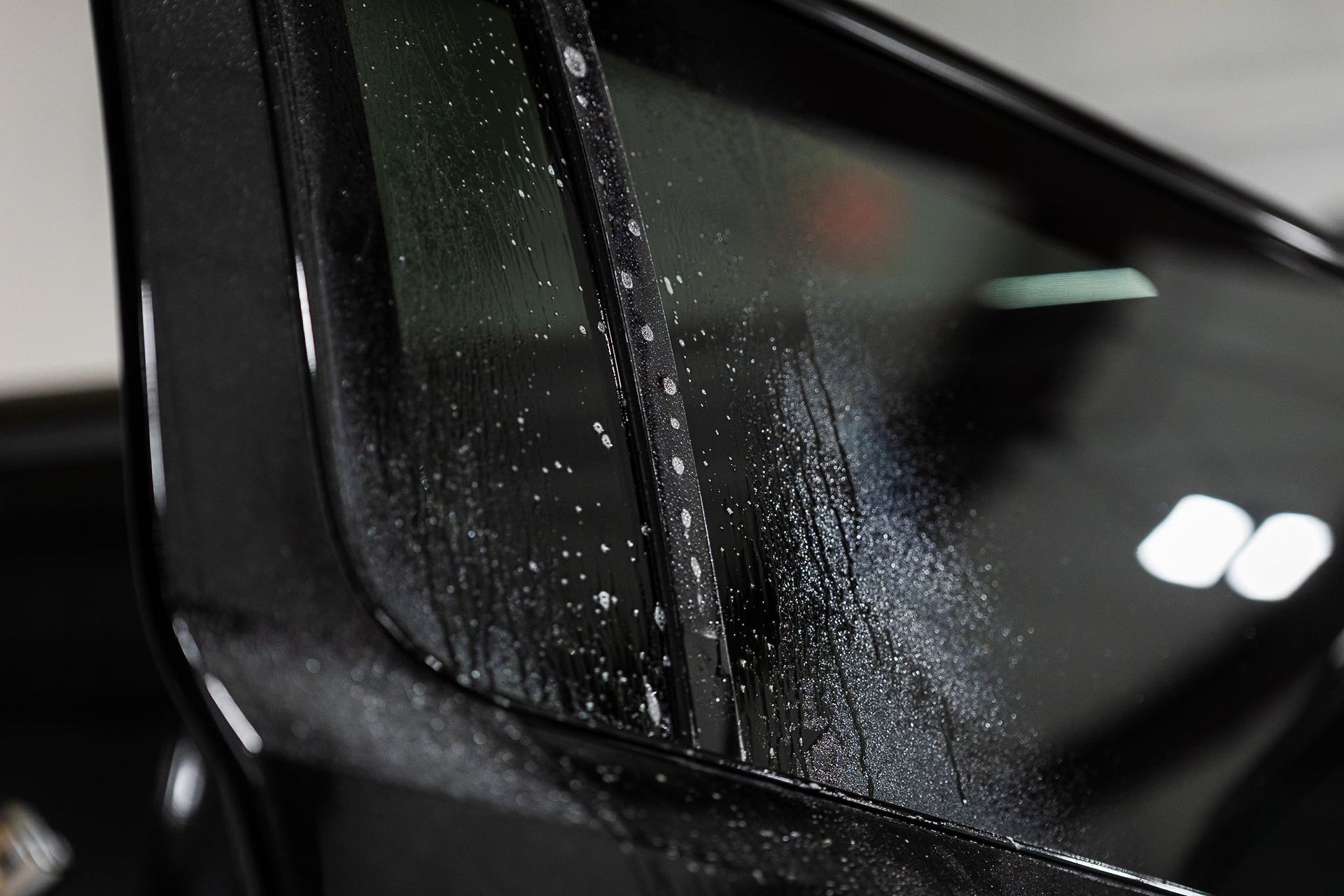 Close-up of a black car window covered in water droplets, with bright reflections visible.