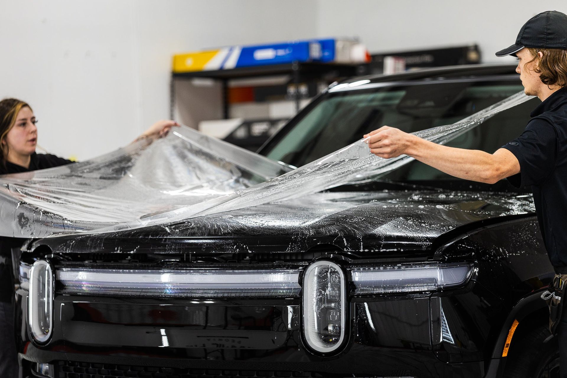 Two people wrapping a black SUV in clear protective film inside a garage