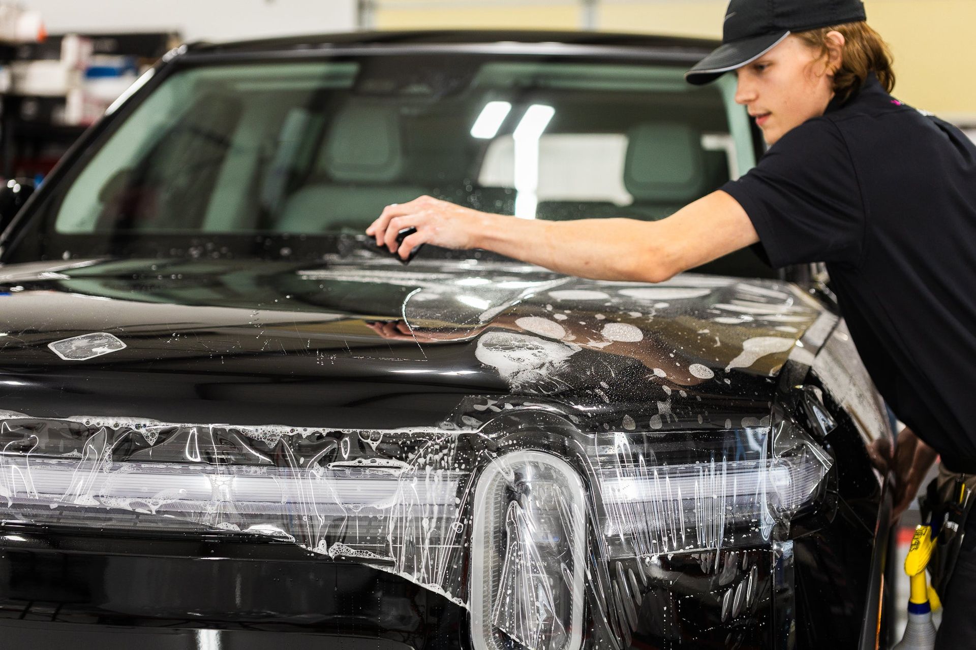 Person polishing a black car hood in a garage, covered in soapy foam