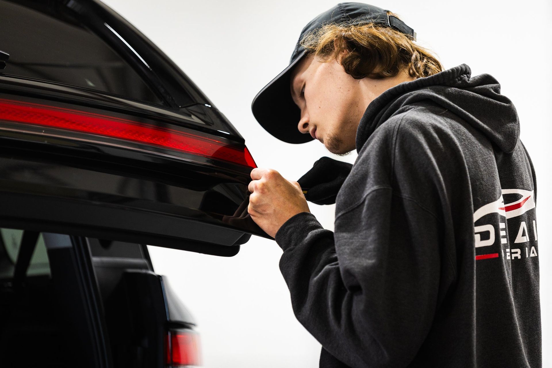 Person opening the trunk of a black car, wearing a dark hoodie and cap