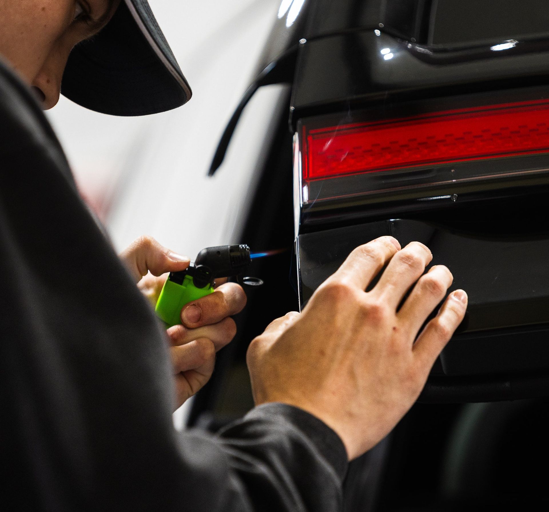 Hands using a green tool to inspect or repair a black car’s rear light area.