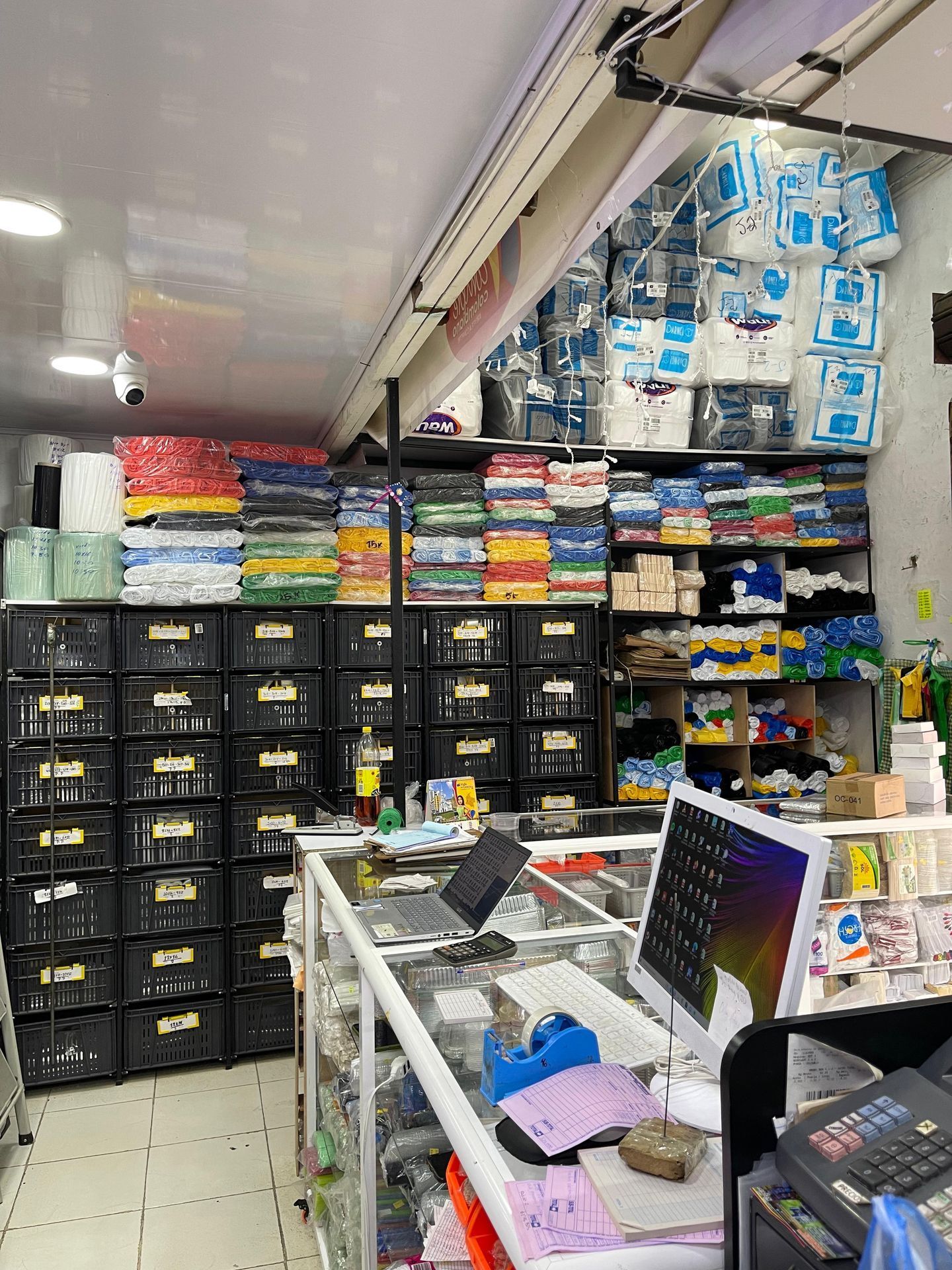 Pasillo de una pequeña tienda con cajas negras apiladas, paquetes coloridos.