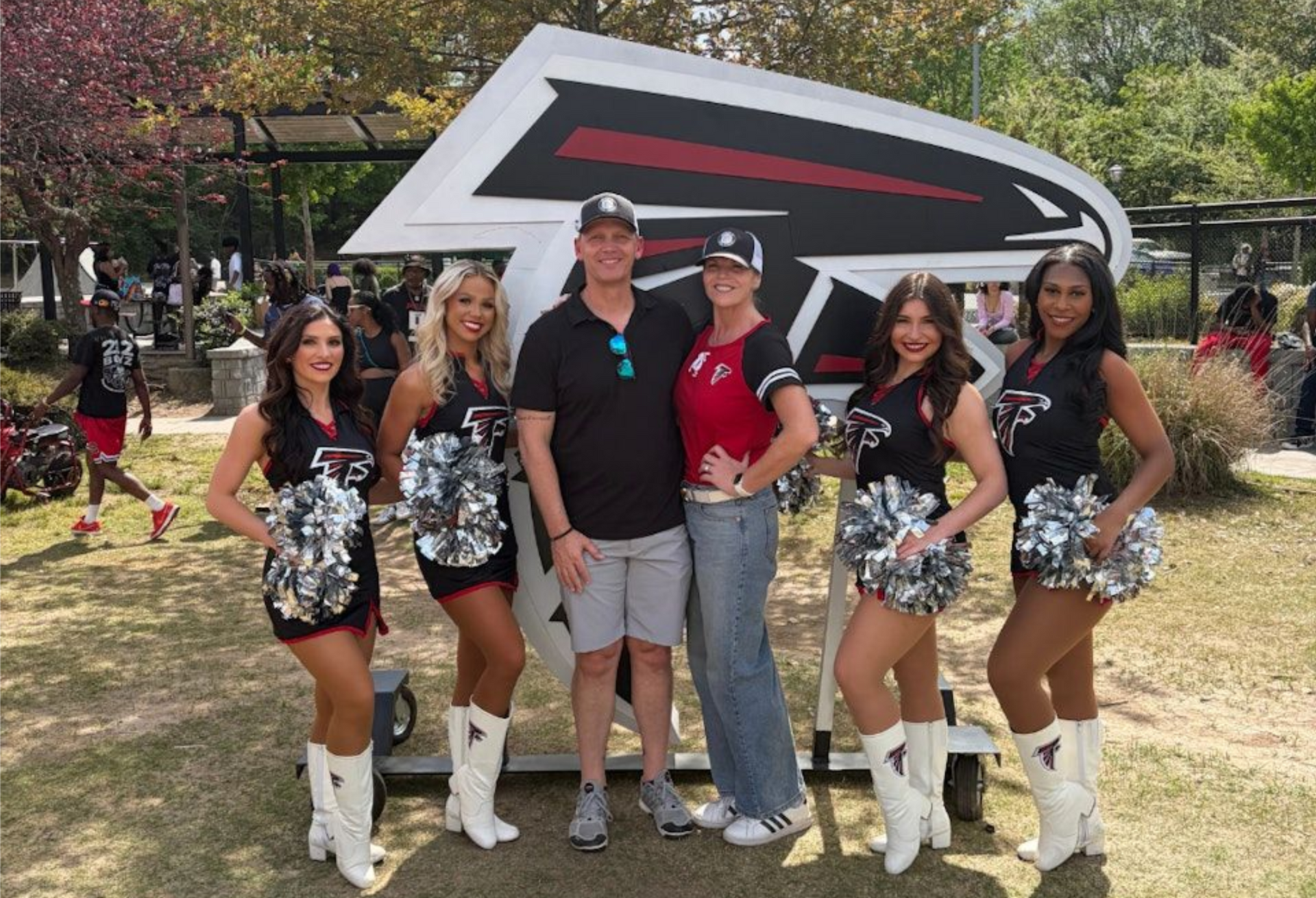 Group posing outdoors with a large hawk logo backdrop, flanked by two women in matching outfits and white boots.