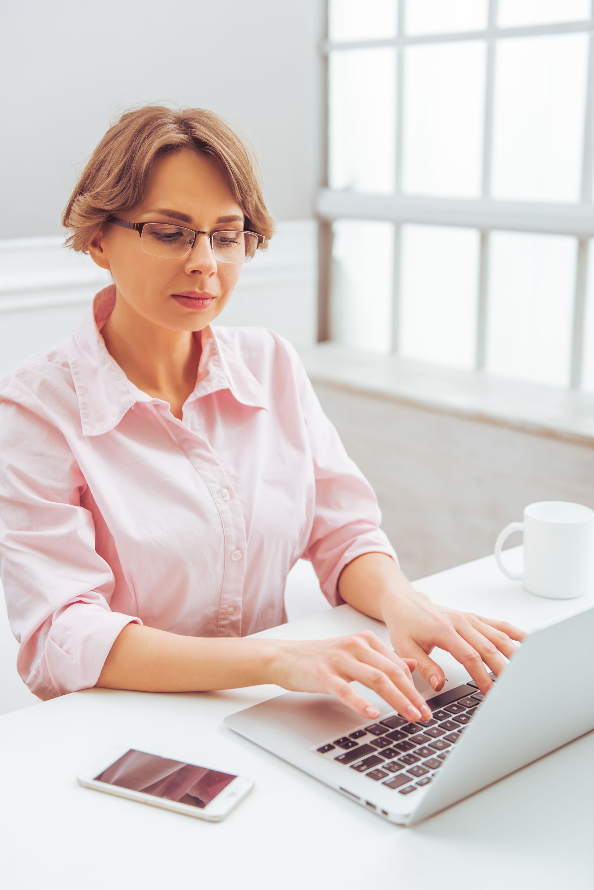 Woman in a pink shirt typing on a laptop at a bright desk with a smartphone and coffee mug.