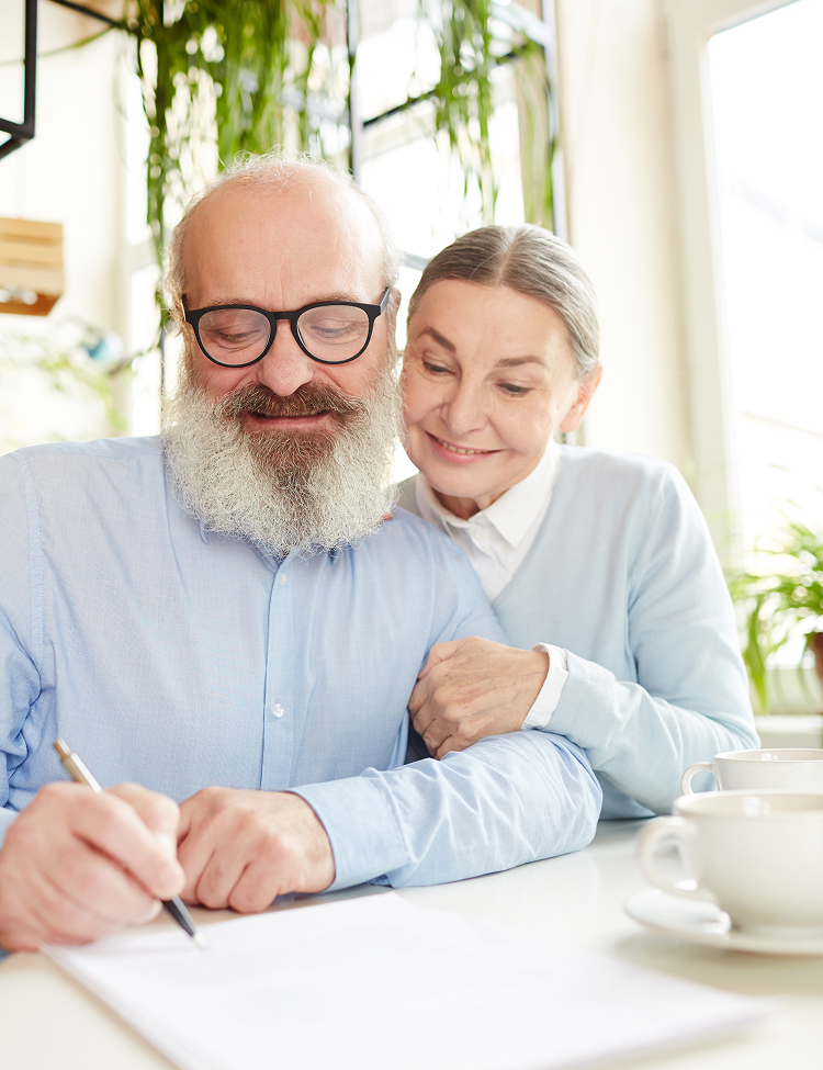 Two people smiling while writing together at a bright table with papers and a cup of coffee