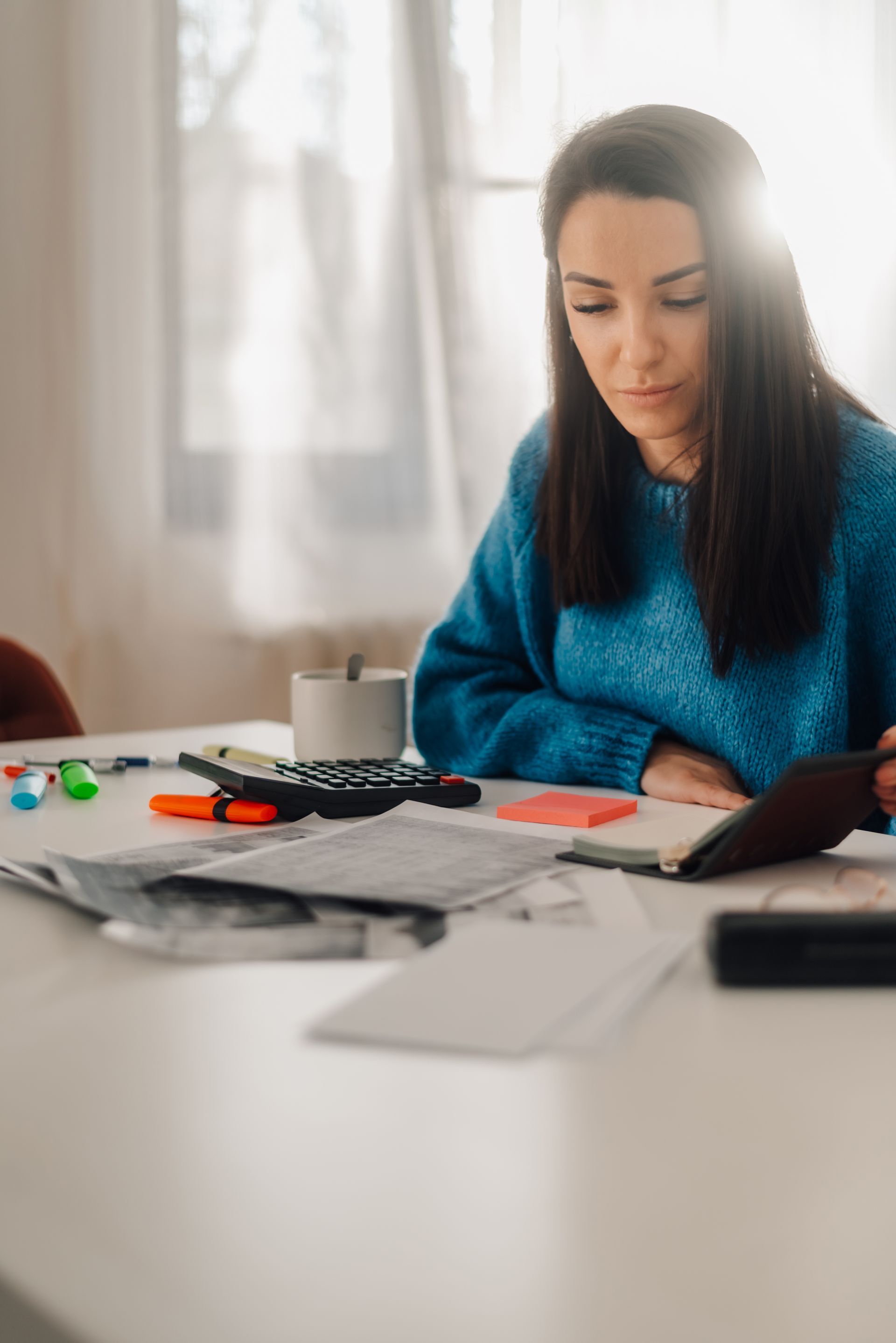 Woman in blue sweater writing at a desk with papers, markers, and a mug near a bright window