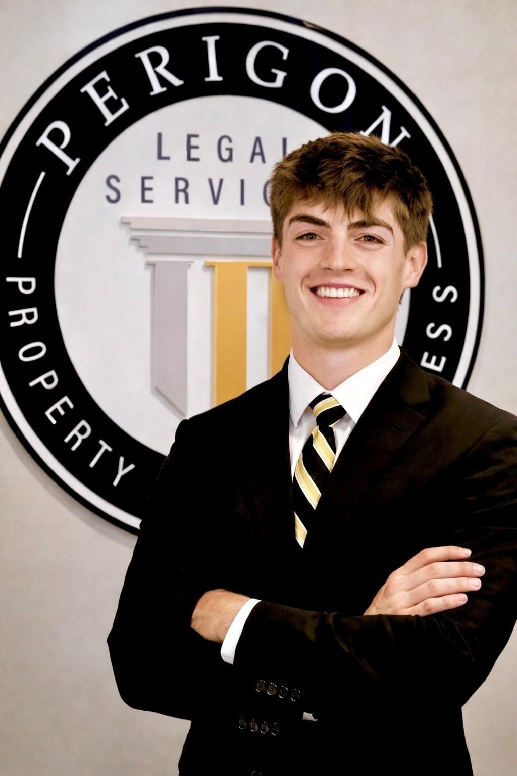 Young man in a black suit smiling with arms crossed in front of a PERIGON Legal Services sign.