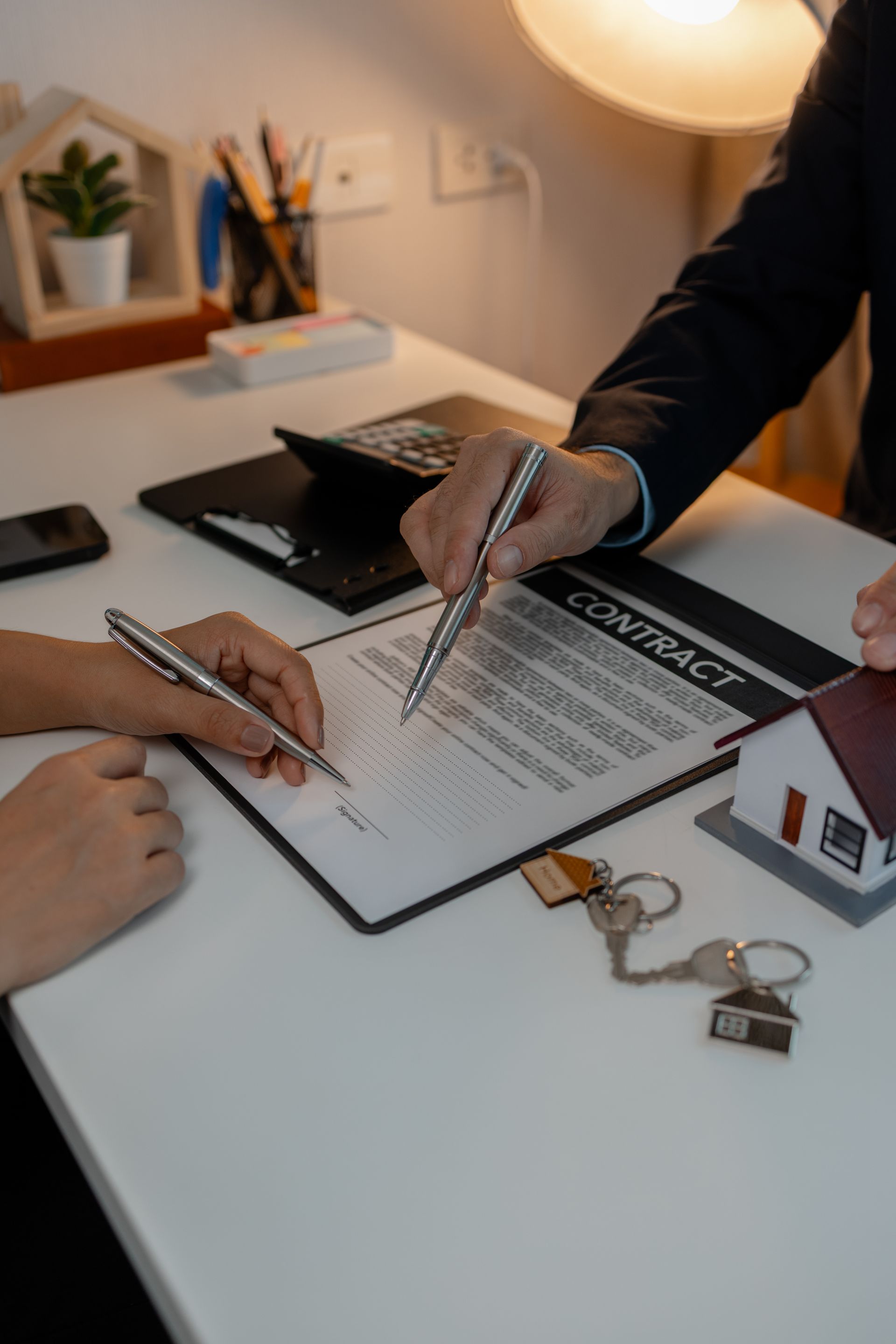 Hands pointing at a signed document on a desk with keys and a small house model in a bright office