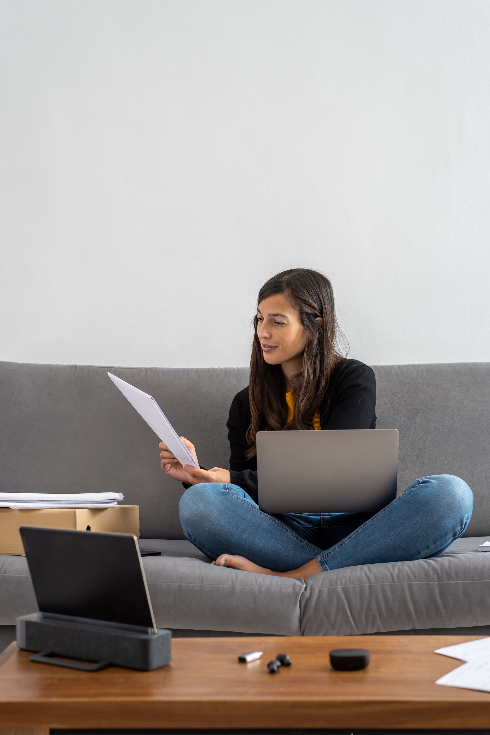 Person sitting cross-legged on a couch with a laptop and papers in a bright living room.