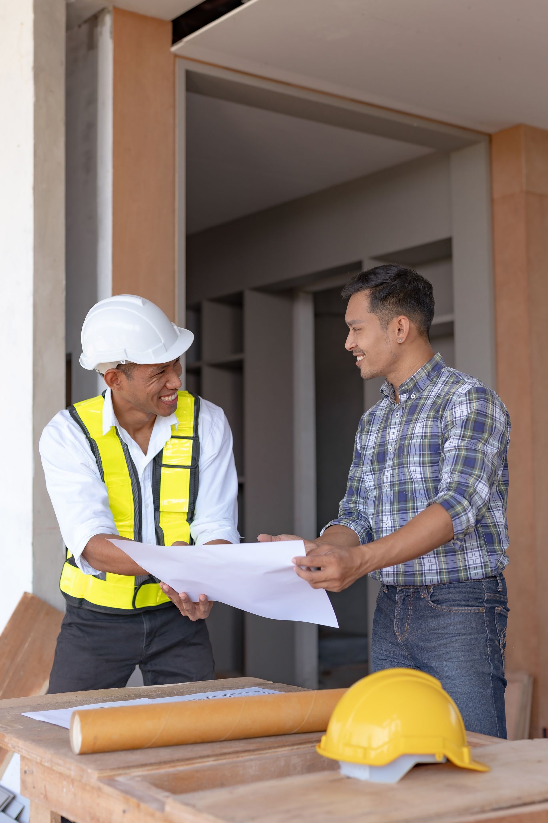 Two construction workers reviewing blueprints at a building site, with a hard hat on the table.