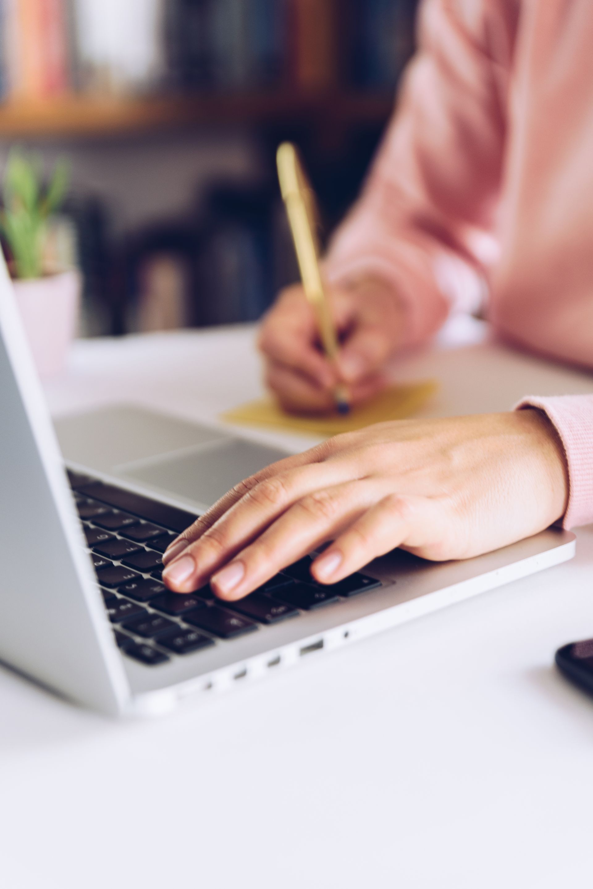 Hands typing on a silver laptop while writing in a notebook at a desk