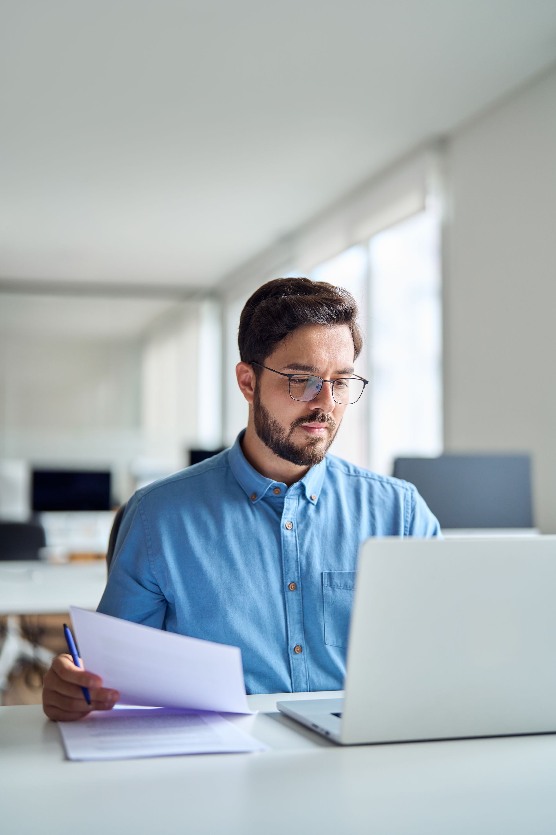 Person working at a laptop while reviewing papers in a bright office