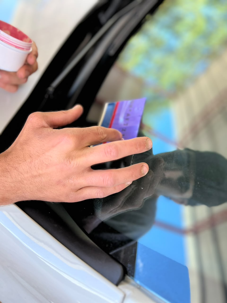 Una mano sostiene una tarjeta morada contra la ventanilla de un coche, con una taza blanca cerca.