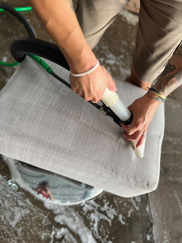 Una persona lava una silla de malla gris con una manguera y un cepillo sobre un suelo mojado.