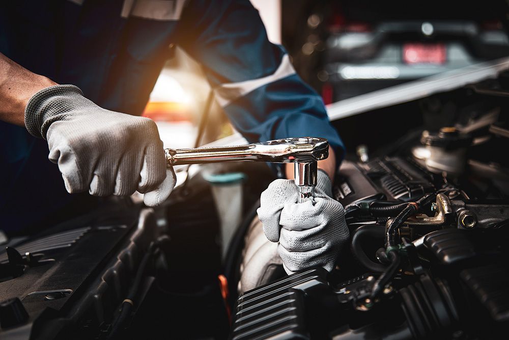 A close-up of hands in work gloves using a ratchet wrench to repair a car engine in a workshop.