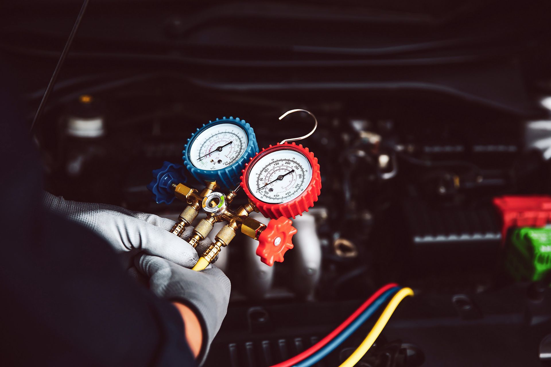 Gloved hands hold an AC manifold gauge set with red and blue dials in front of an open car engine bay.
