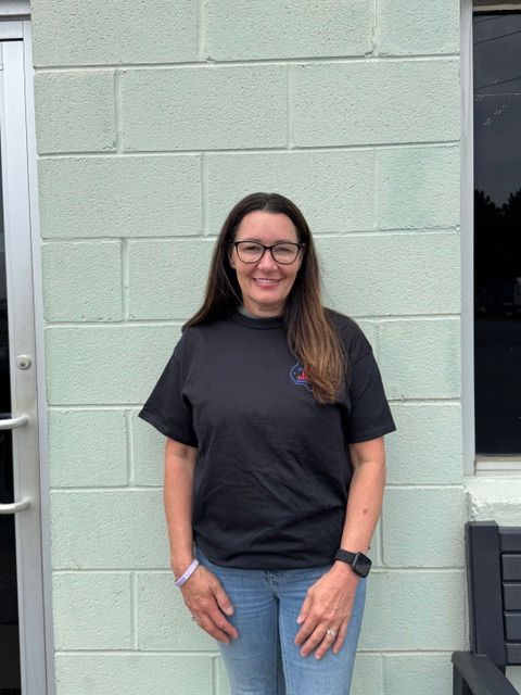 Woman standing against a light green brick wall, wearing a black T-shirt, blue jeans, and glasses, smiling.