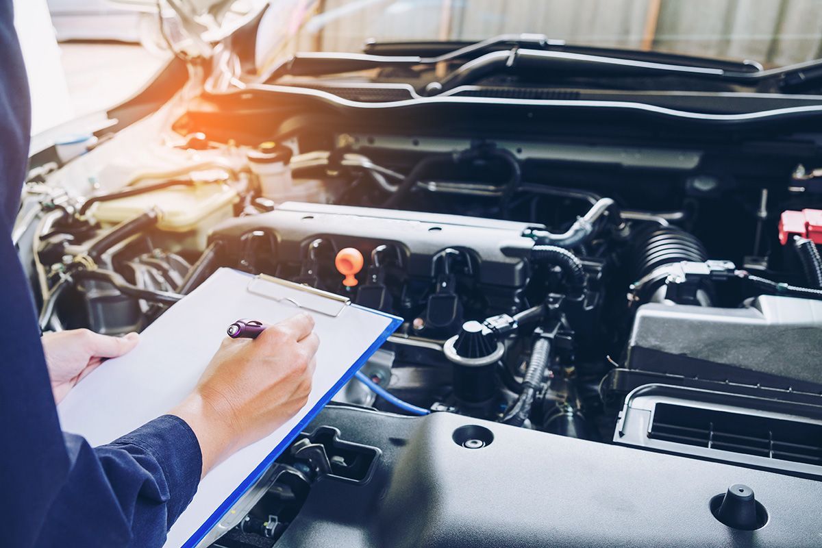 A mechanic wearing a blue uniform holds a clipboard while inspecting an open car engine.