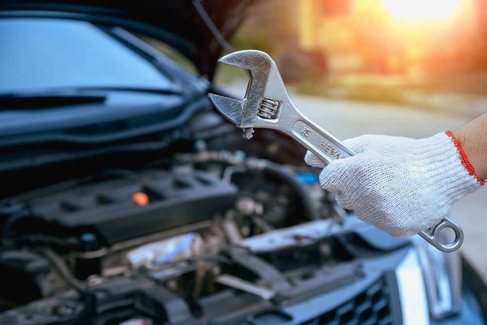 A hand in a white work glove holds an adjustable wrench over the open engine bay of a car, illuminated by sunlight.