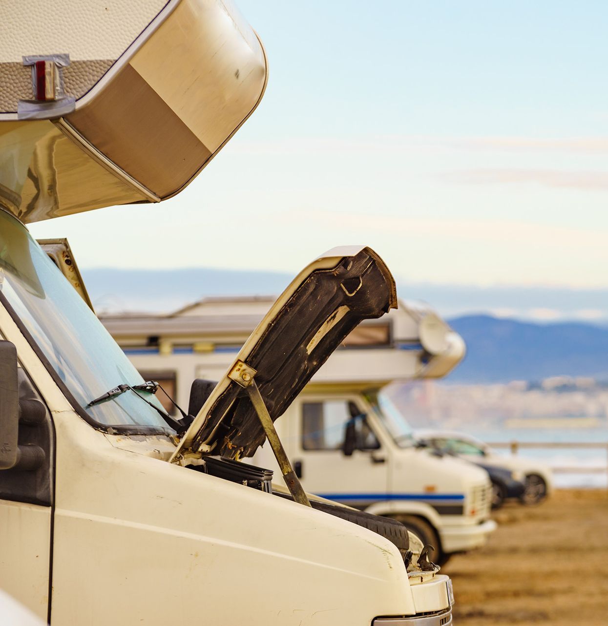 A light-colored camper van with its hood popped open, parked in a lot by the coast with another camper van in the back.