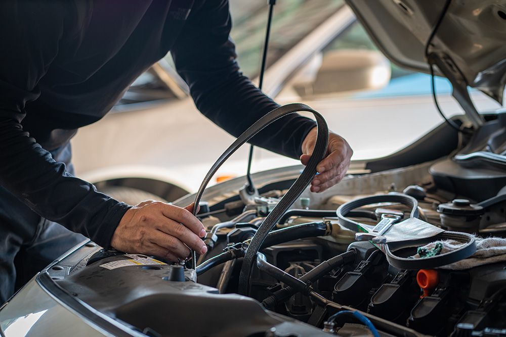A mechanic holding a serpentine belt while working on an open car engine in a garage.