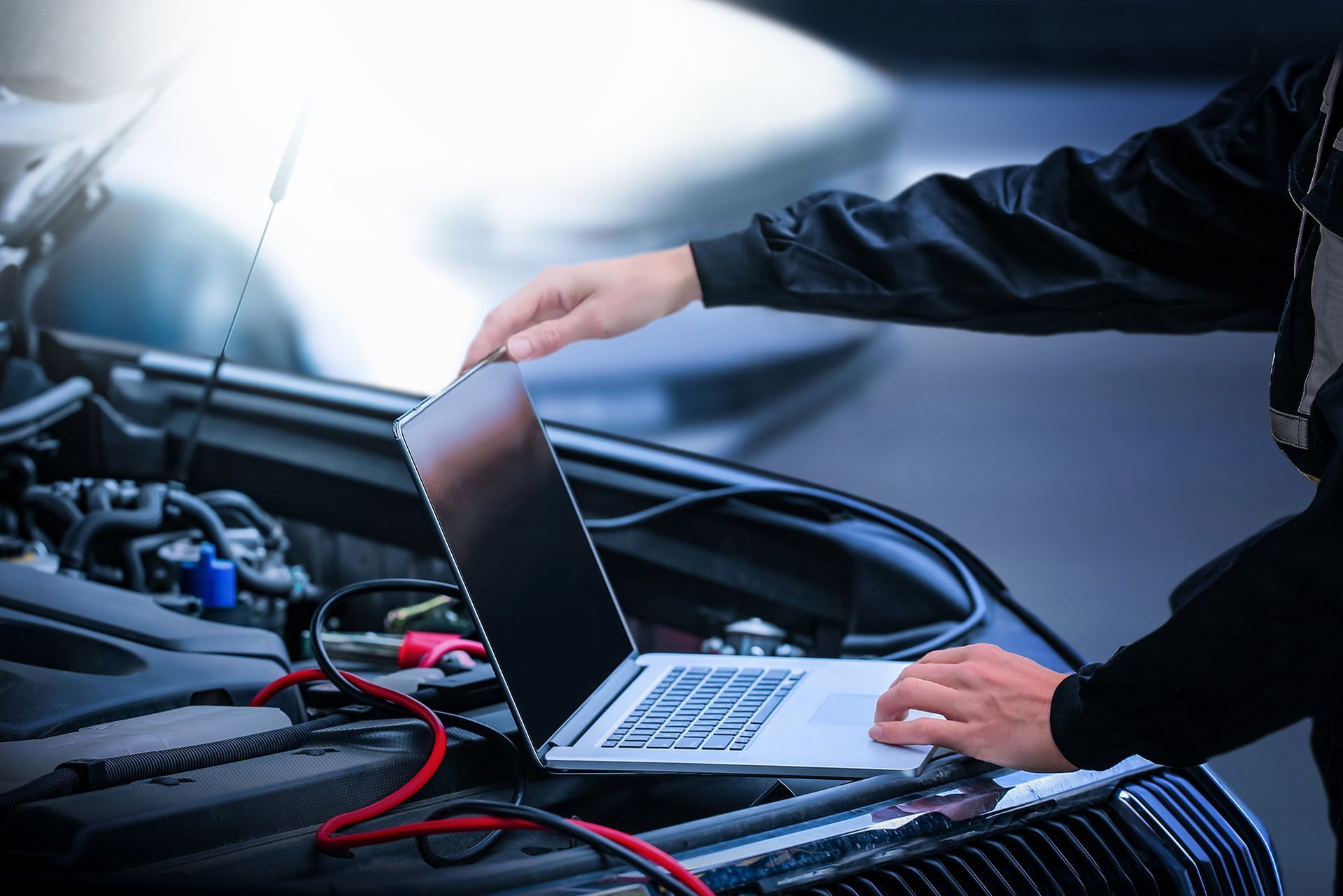 A mechanic uses a laptop connected to a car engine with jumper cables to perform a diagnostic test.