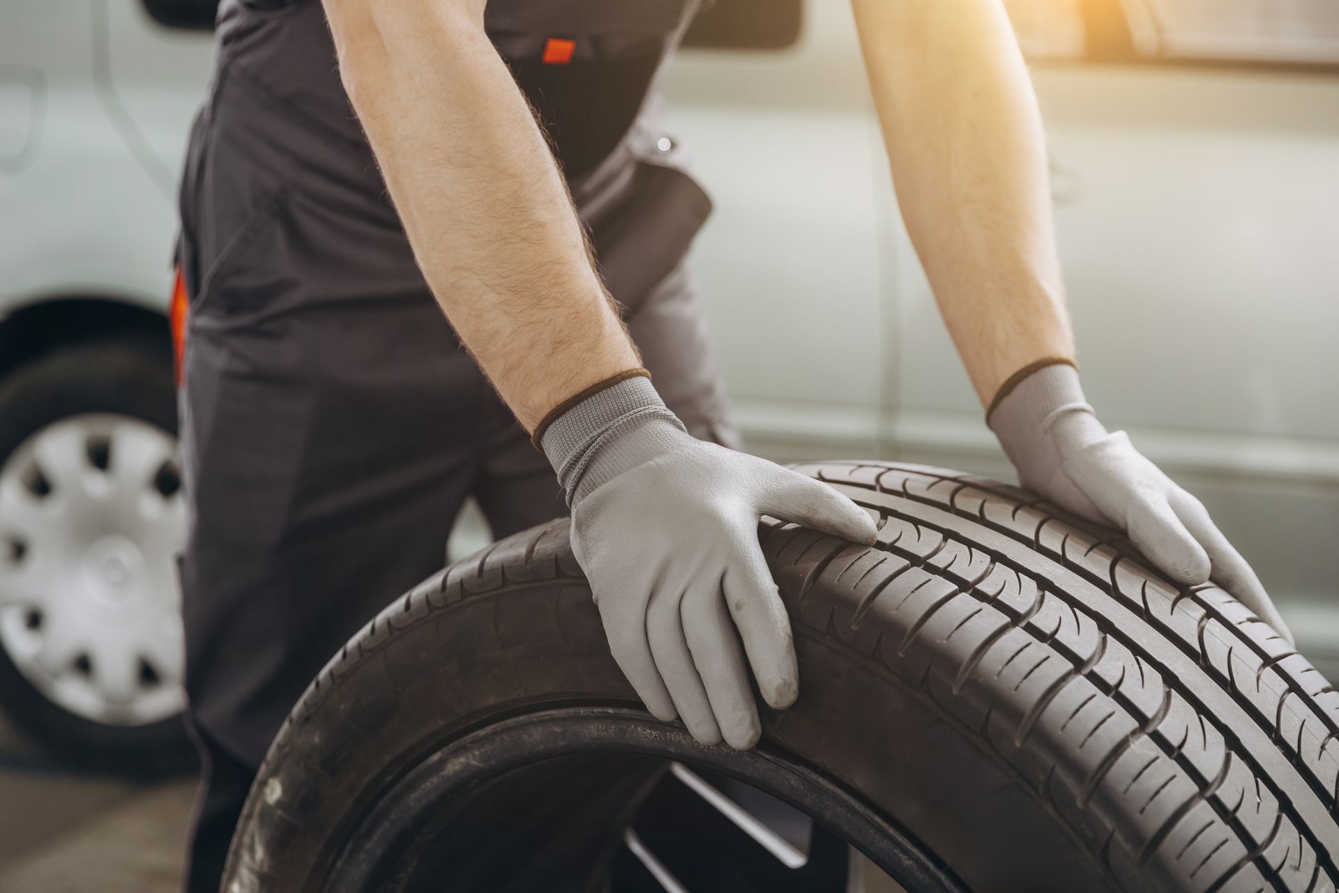 A mechanic wearing gray work gloves holds a car tire in an auto repair shop.
