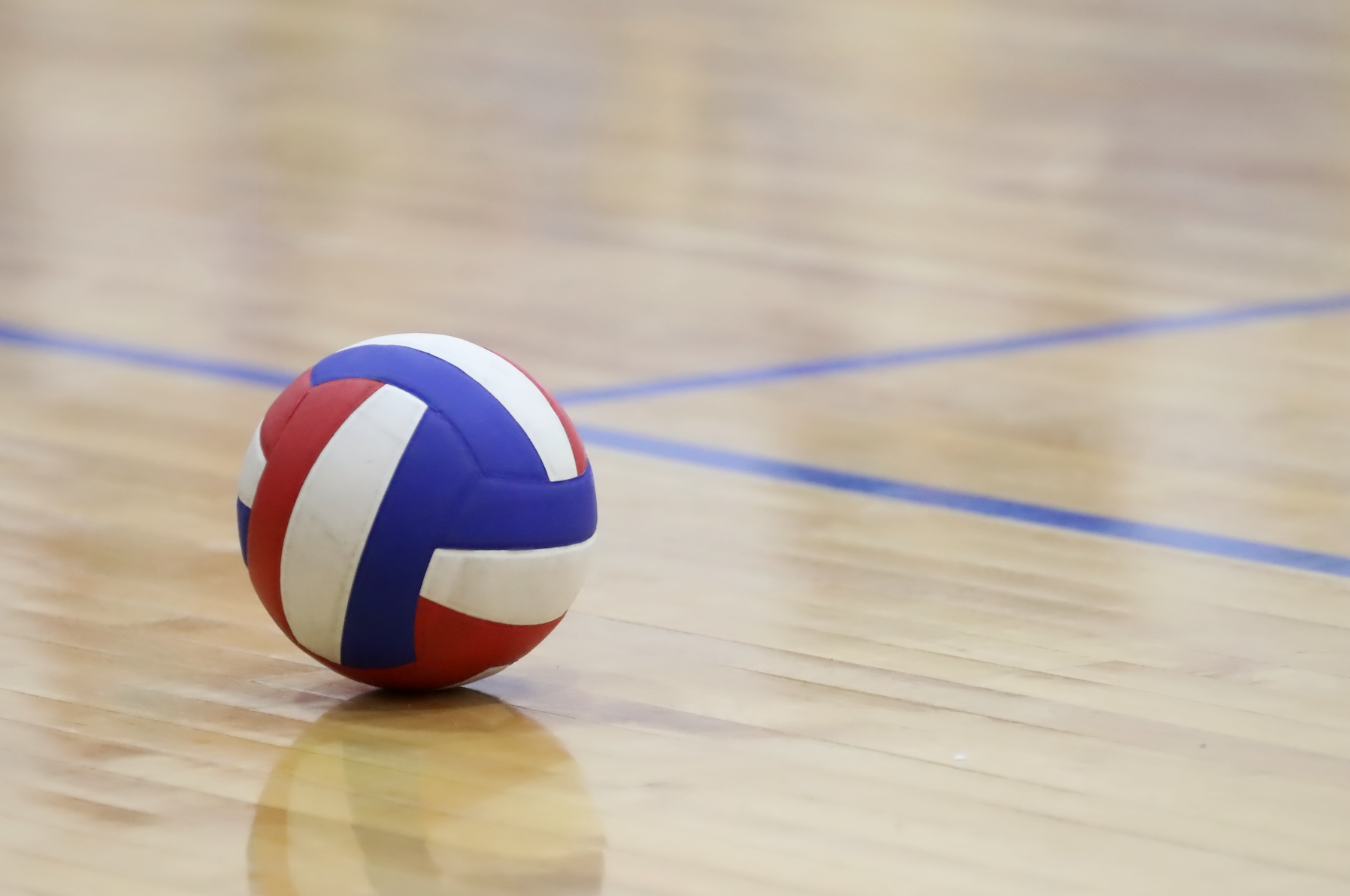 A red , white and blue volleyball is sitting on a wooden court.