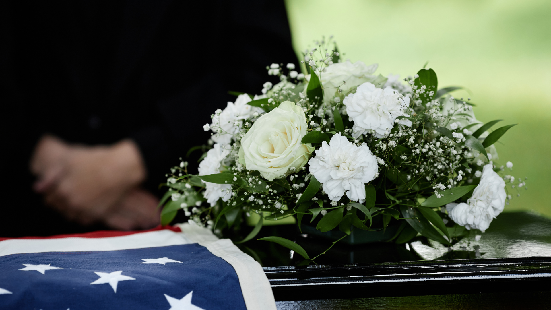 American flags stand at tombstones in a cemetery, with a tree and flowers in the foreground.