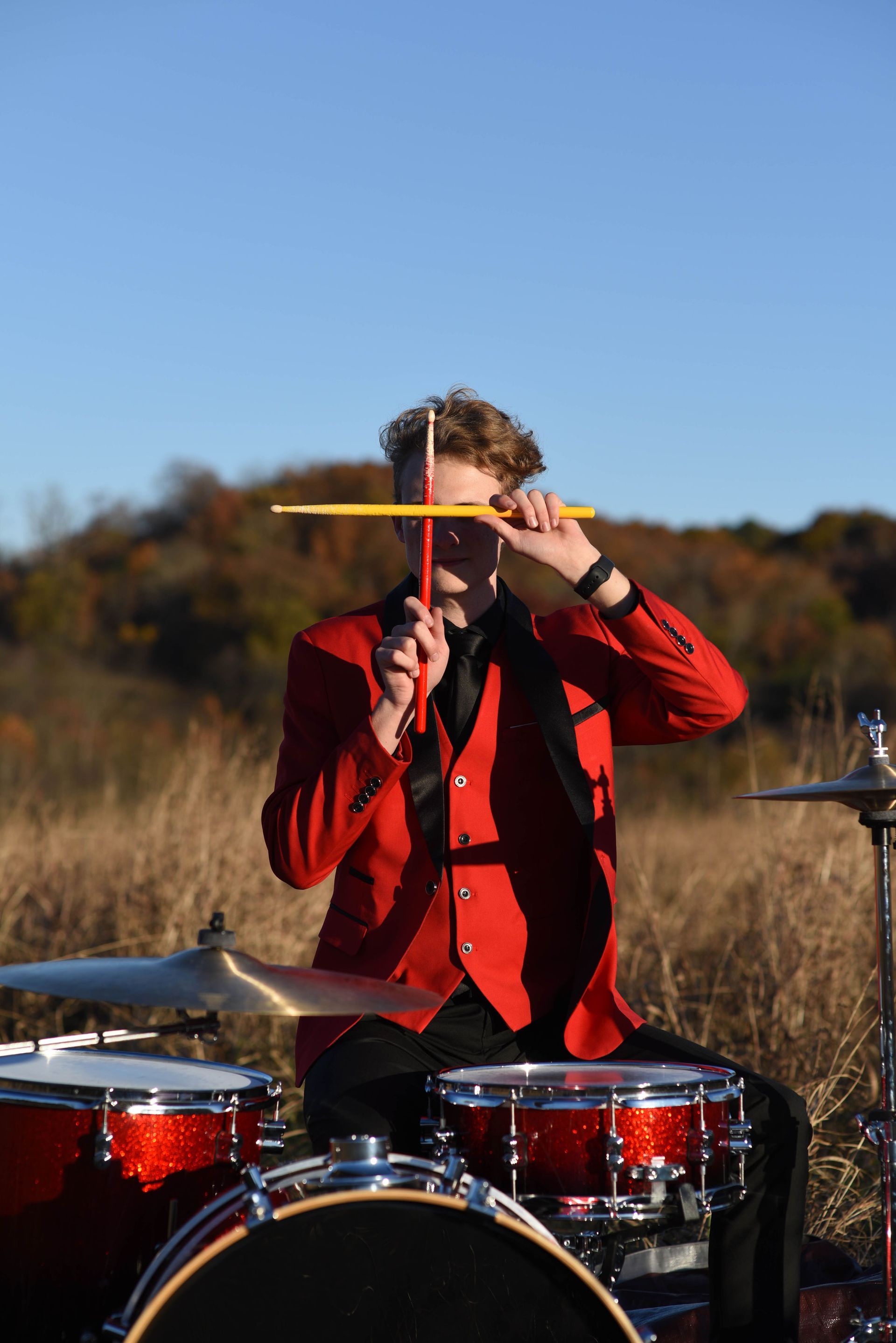 Drummer in a red jacket performing outdoors behind a drum kit under a blue sky