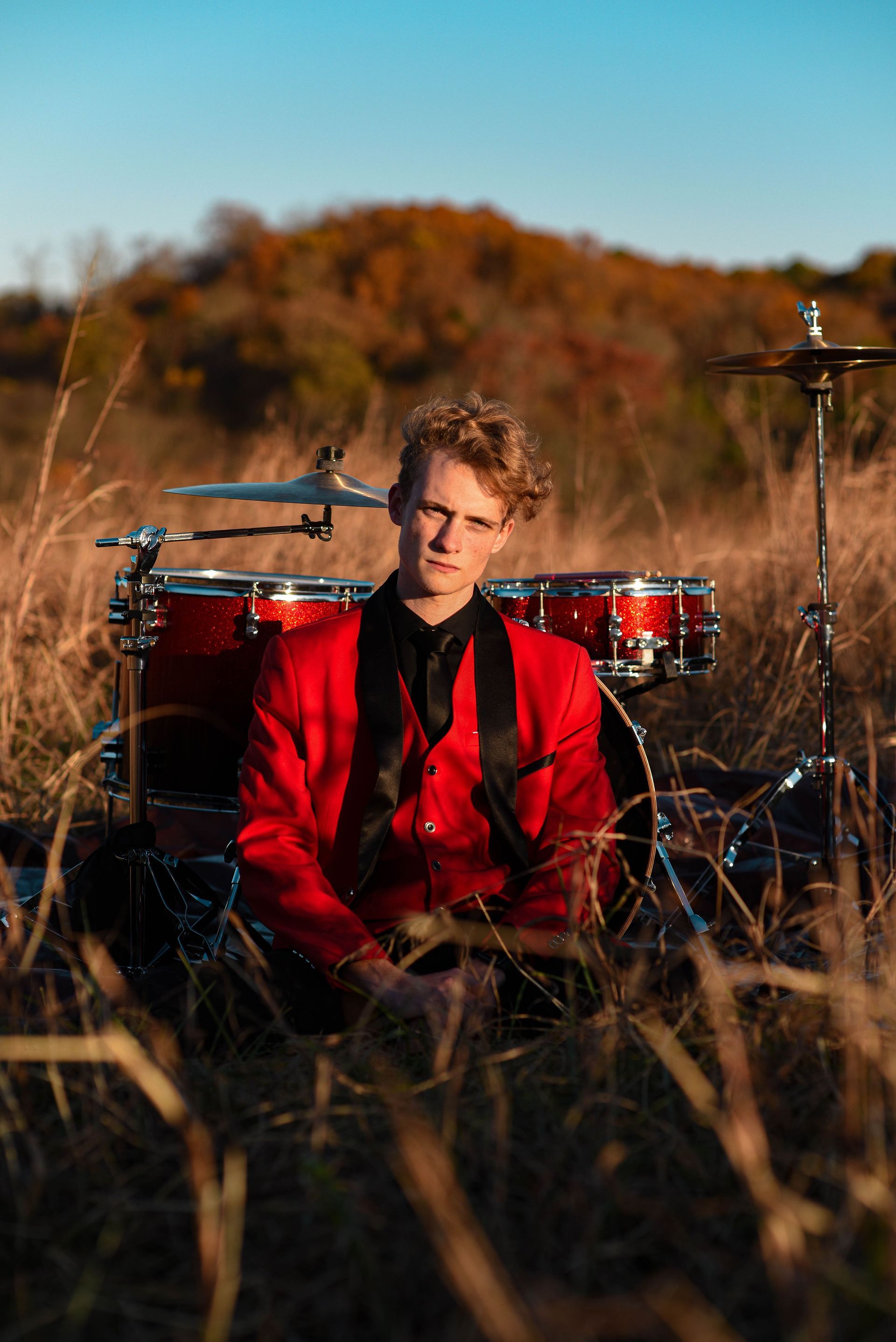 Person in a red jacket sitting in tall grass, with a red drum kit and cymbal in the background.