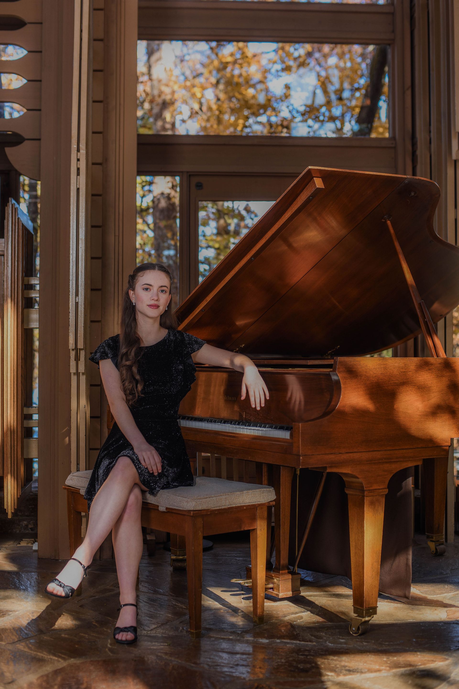 Woman seated beside a grand piano in a sunlit room with large windows and autumn trees outside