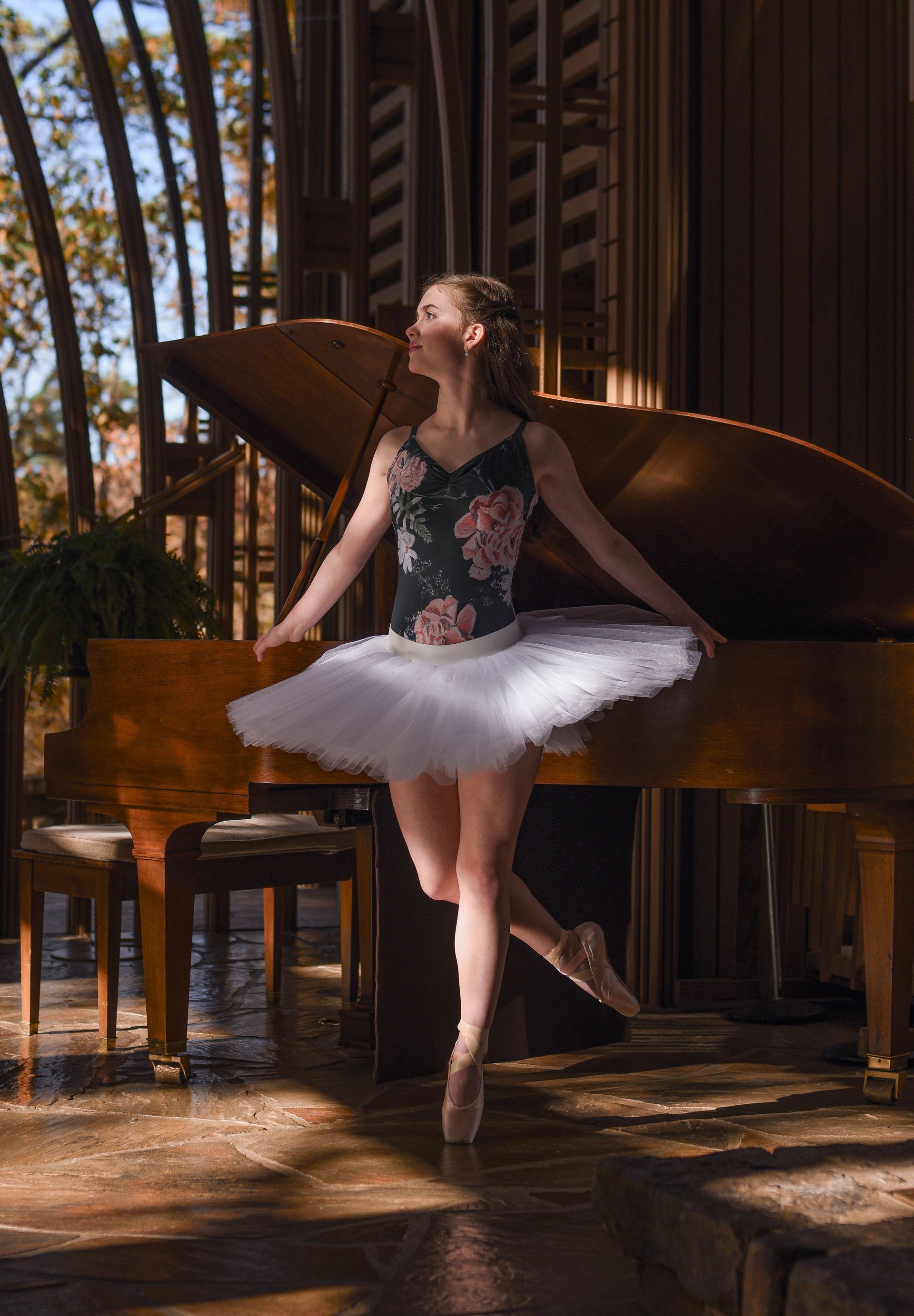 Ballerina in a white tutu posing beside a grand piano in a sunlit rustic hall