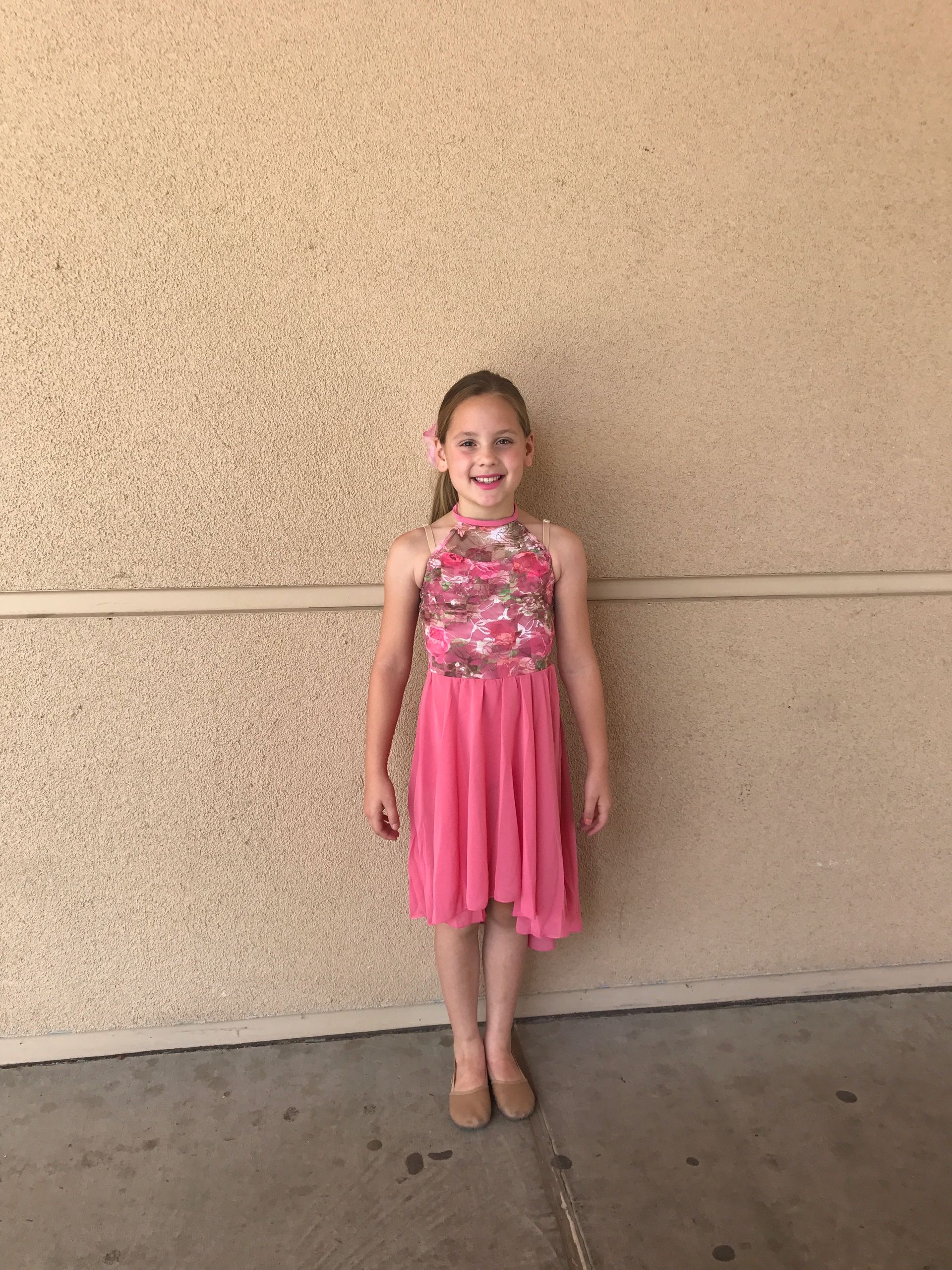 Girl in a pink dress standing and smiling against a beige wall.